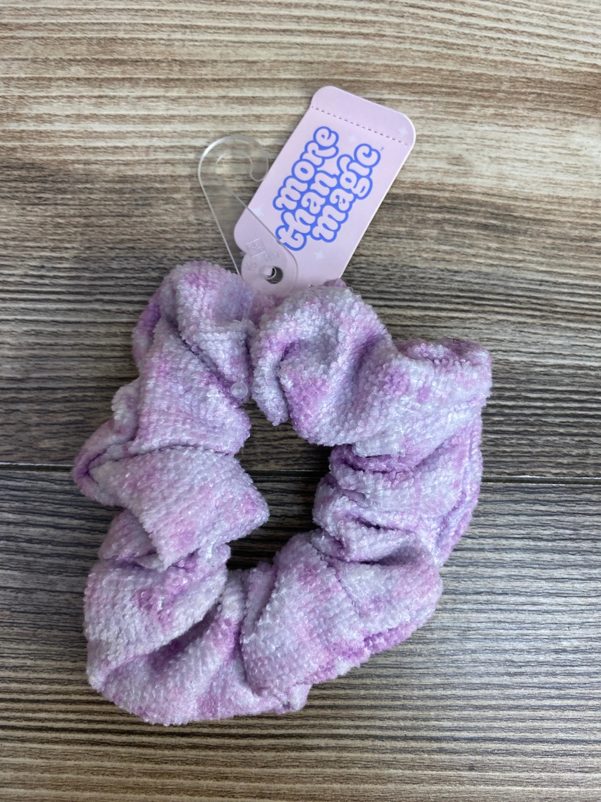 Purple and white textured fabric scrunchie with 'more than magic' tag on wood surface