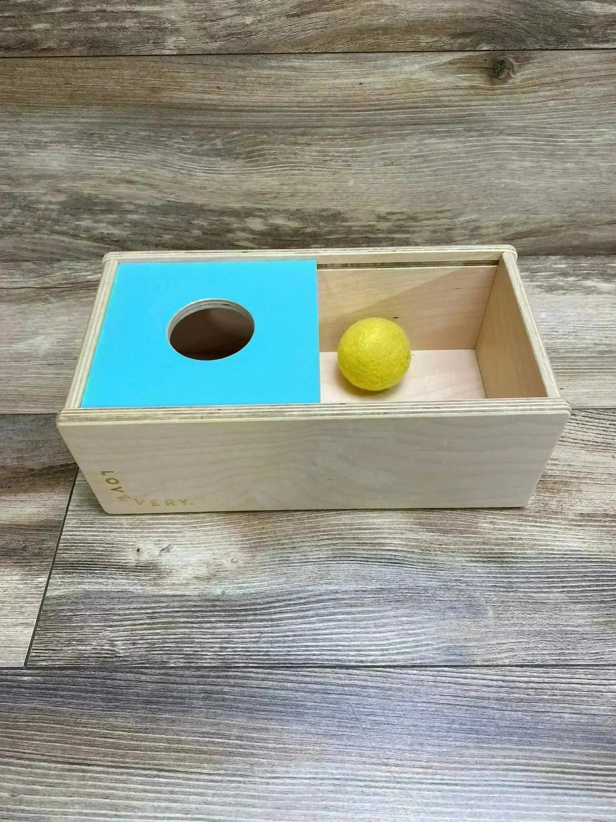 Angled top view of Lovevery Sliding Top Box showing turquoise lid with circular hole and yellow ball on wood backdrop