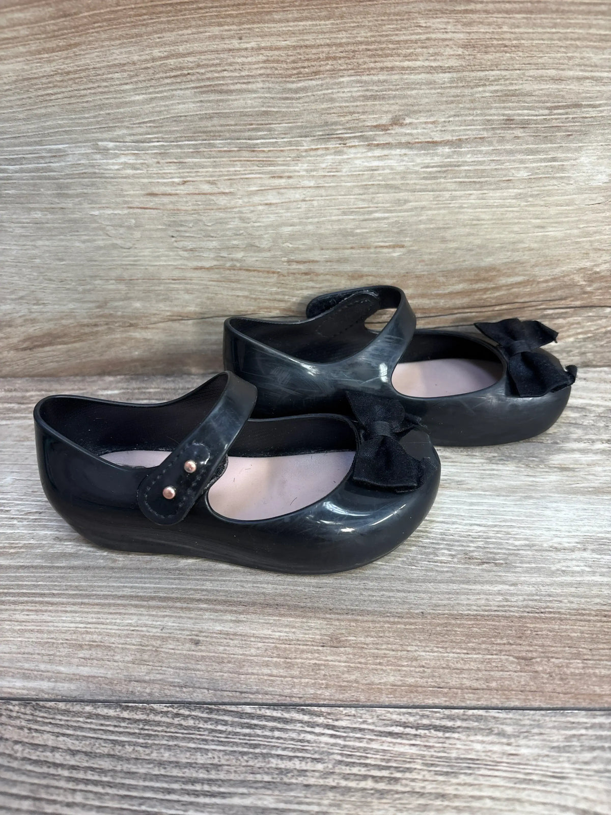 Side view of Mini Melissa Ultragirl black jelly Mary Jane flats showing bow detail, strap snaps, and pink insoles on wooden background