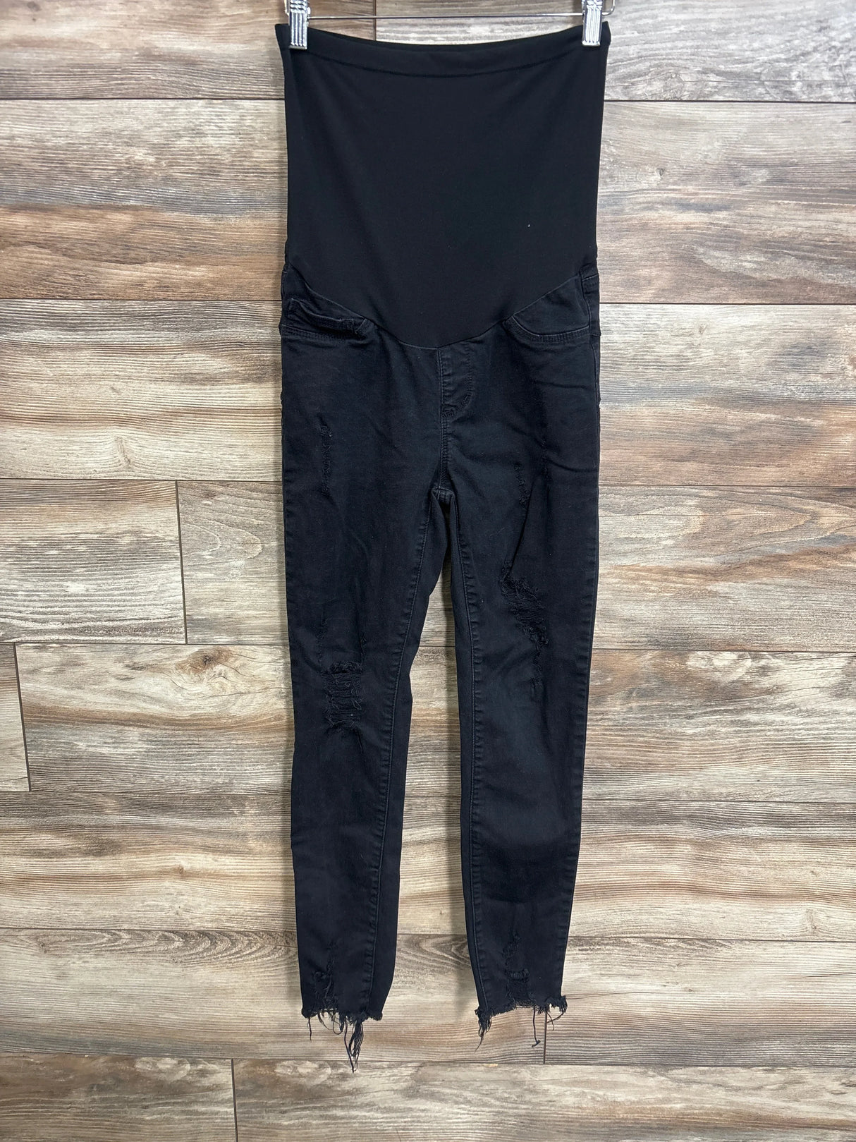 Front view of black SONG maternity full-panel distressed jeans showing high elastic belly panel, ripped knees and frayed ankle hems on wood background