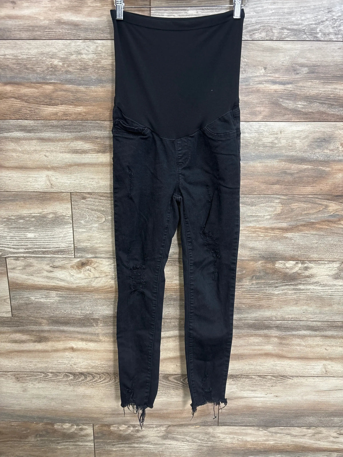 Front view of black SONG maternity full-panel distressed jeans showing high elastic belly panel, ripped knees and frayed ankle hems on wood background