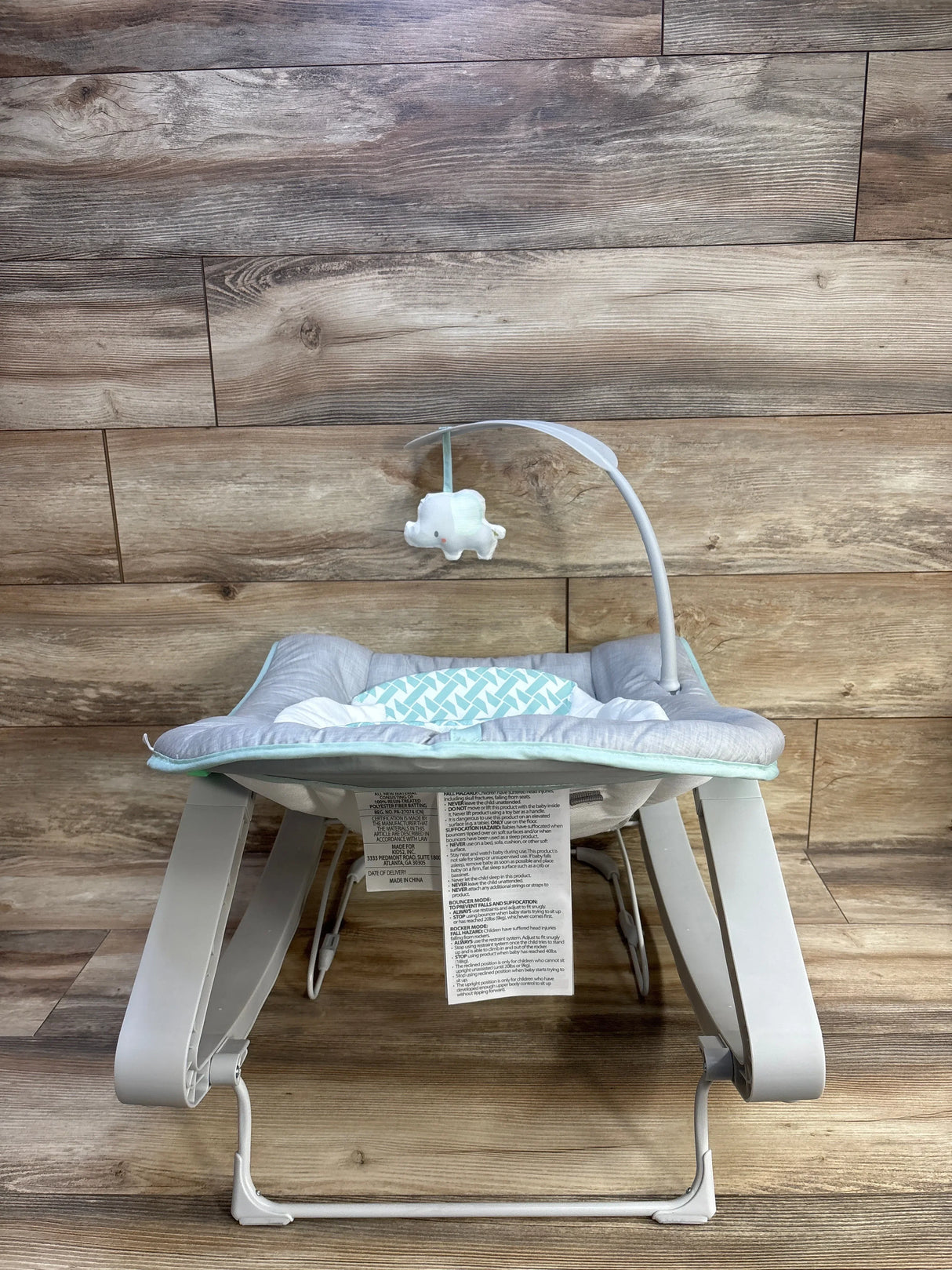 Front view of Ingenuity Keep Cozy bouncer showing padded gray and mint seat, toy elephant arm, and rocker legs on wood floor