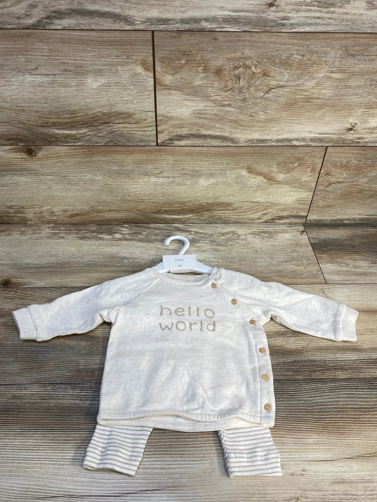 Front flat lay of taupe H&M 3pc baby shirt set size 3M showing long-sleeve top with side snap buttons and 'hello world' text, with striped pants on wood floor