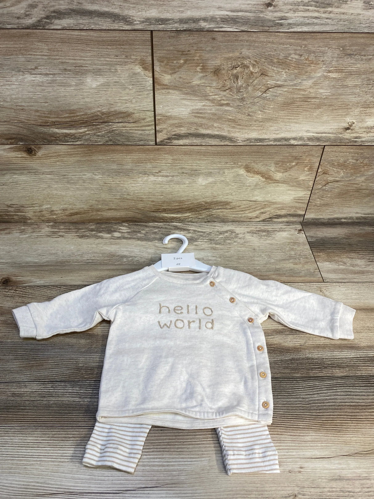 Front flat lay of taupe H&M 3pc baby shirt set size 3M showing long-sleeve top with side snap buttons and 'hello world' text, with striped pants on wood floor