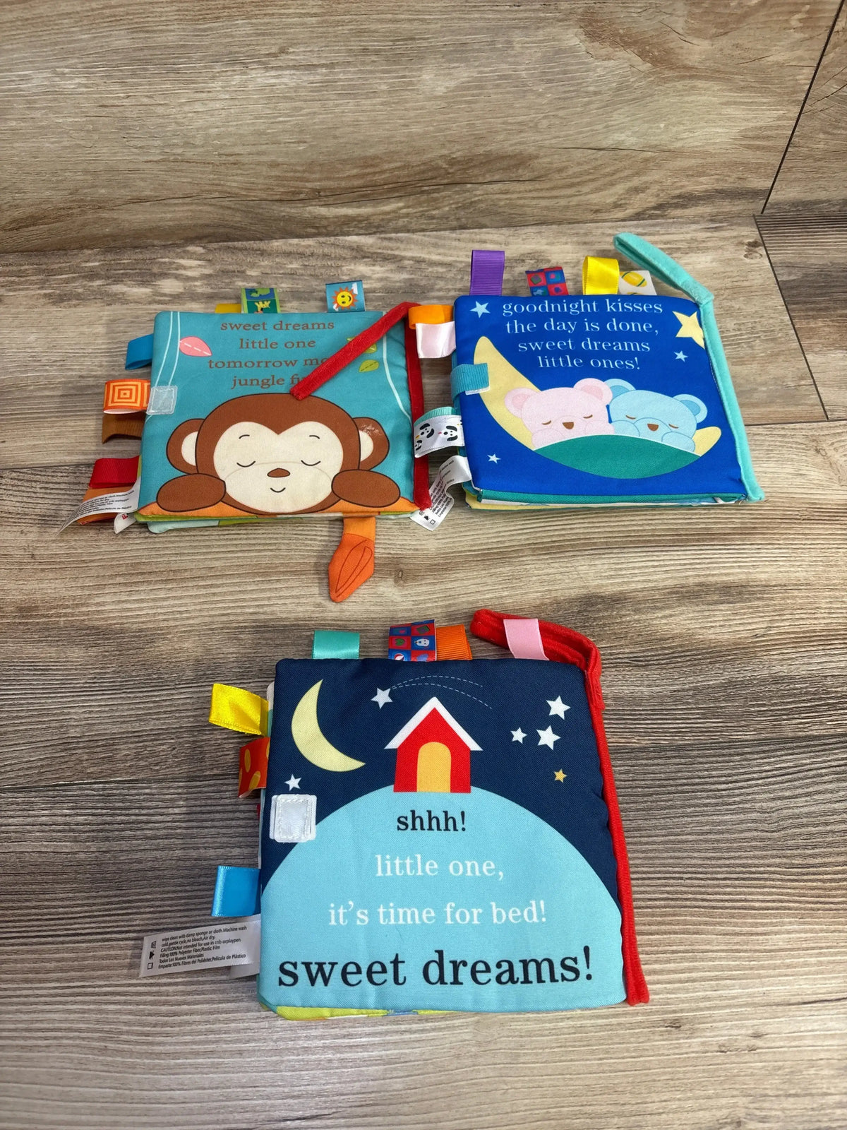 Top view of Funny Zoo Soft Tag Books 3pk showcasing bedtime-themed fabric covers with ribbon tags on a wooden floor