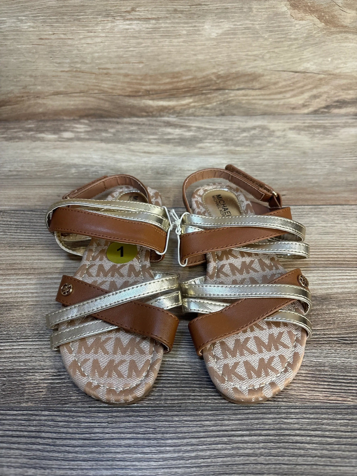 Brown and gold toddler sandals with MK logo print on wood background