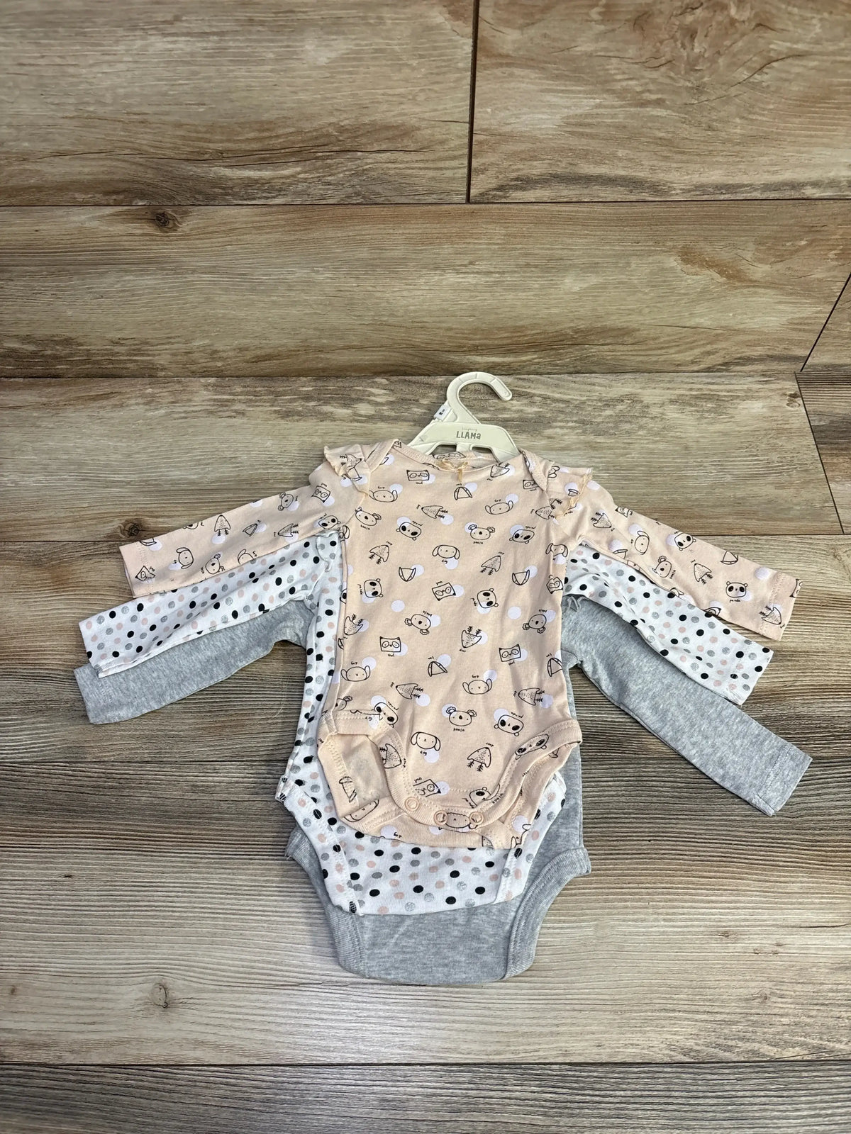 Stack of three long-sleeve baby bodysuits on wood floor, featuring animal prints, polka dots, and solid gray