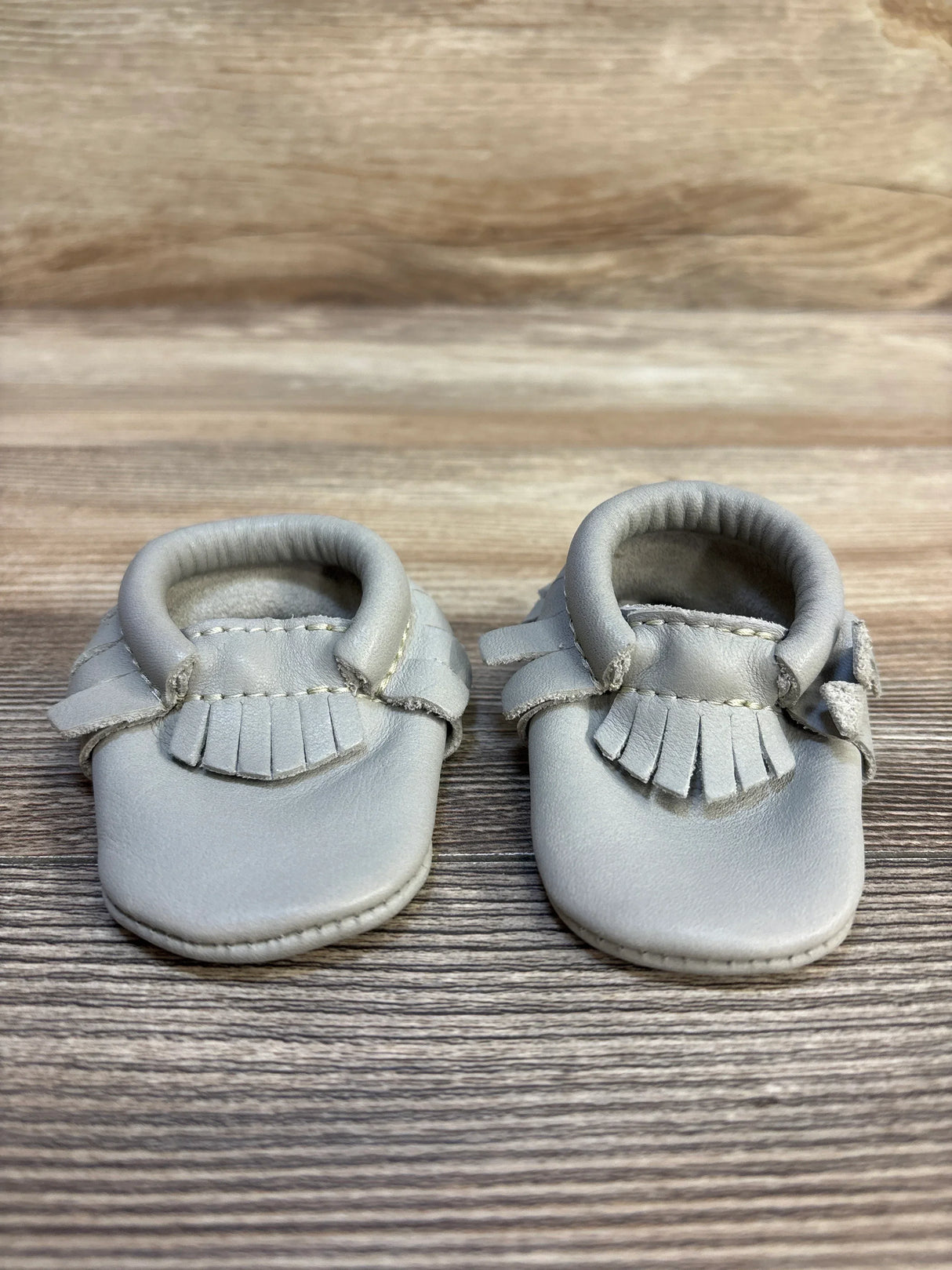 Gray leather baby moccasins with fringe trim on a wooden background