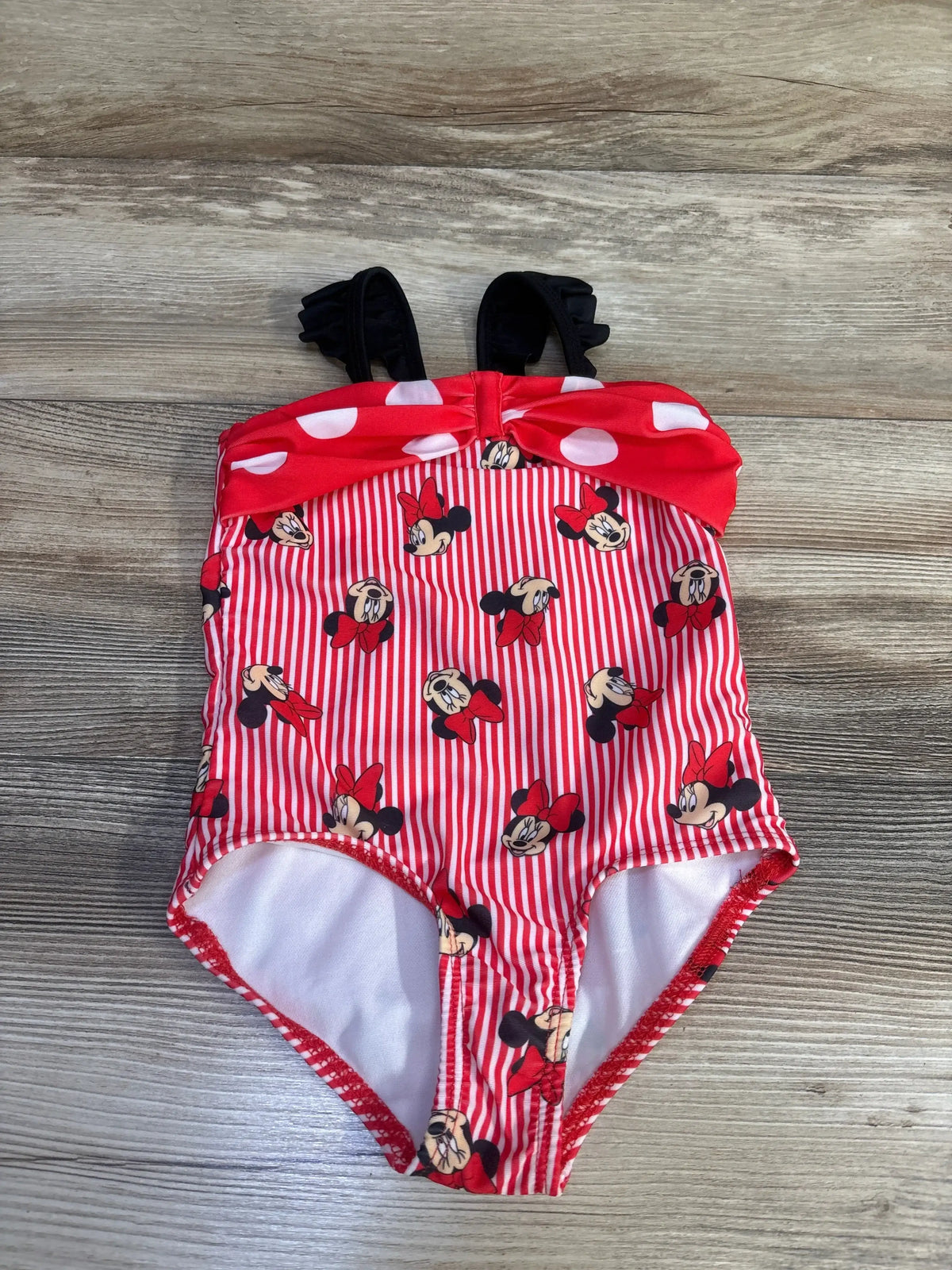 Flat-lay red girls' one-piece Minnie swimsuit with white stripes, Minnie print, polka-dot bow and black ruffle straps on wood