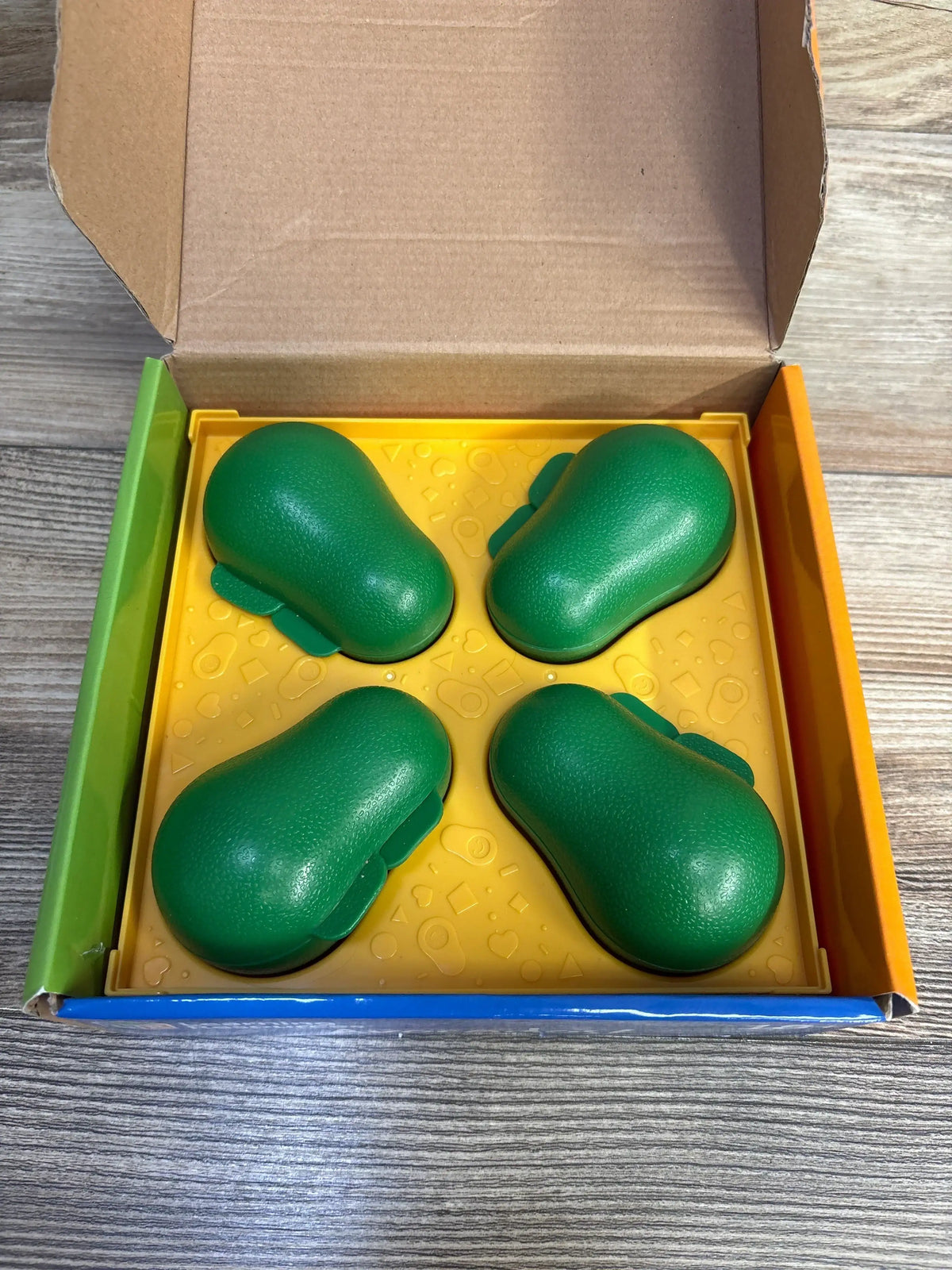 Top view of Learn-A-Lot Avocados 9pc showing four green plastic avocado pieces in a yellow tray inside open box on wood