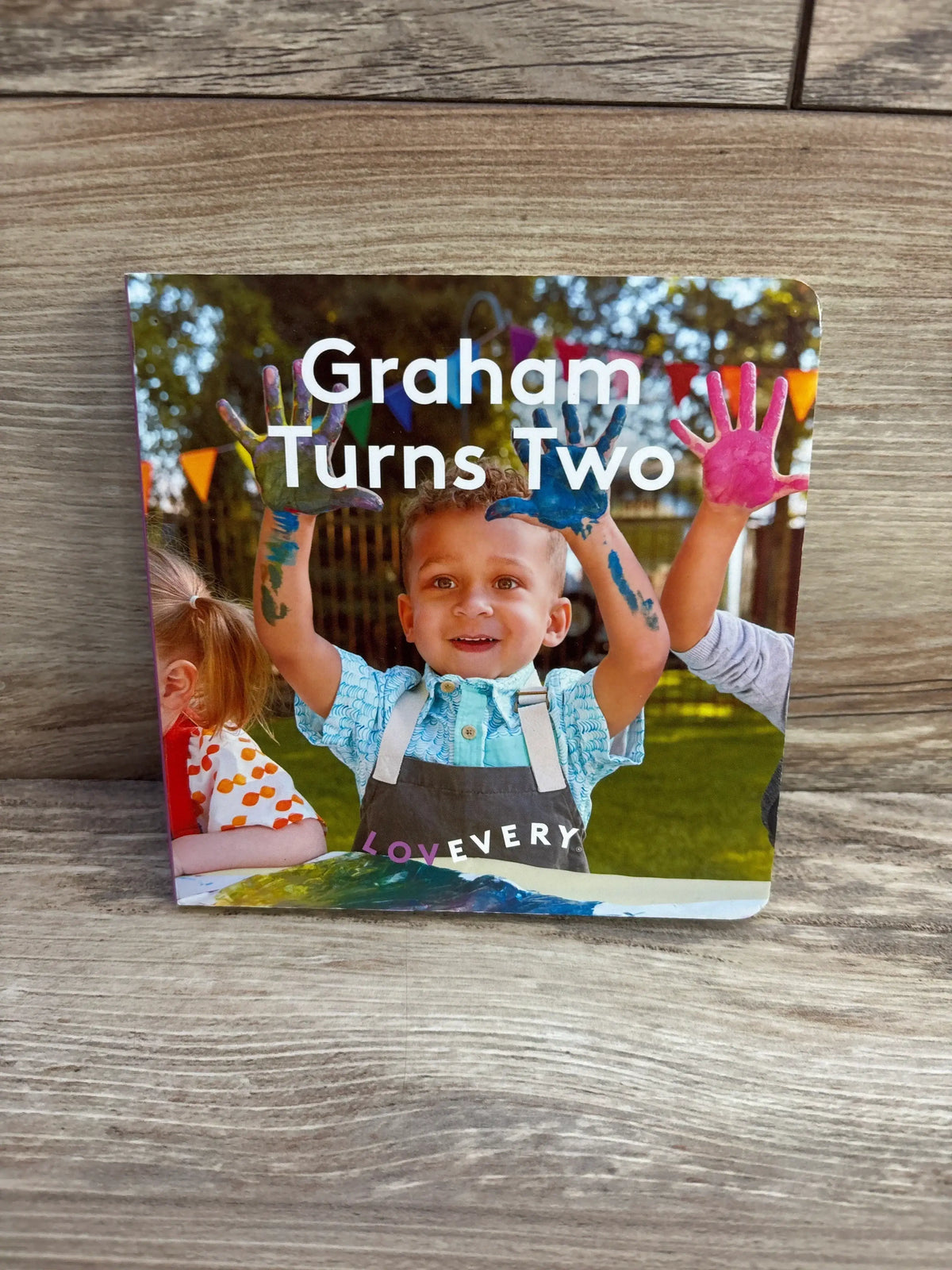 Front view of Lovevery Graham Turns Two board book showcasing the cover photo of a toddler with paint-covered hands with a wooden background