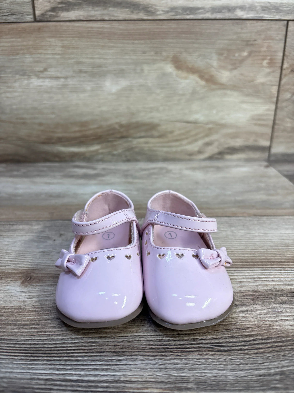 Front view of Teeny Toes patent Mary Jane baby flats in pink showing heart cutouts and bows on a wood plank background