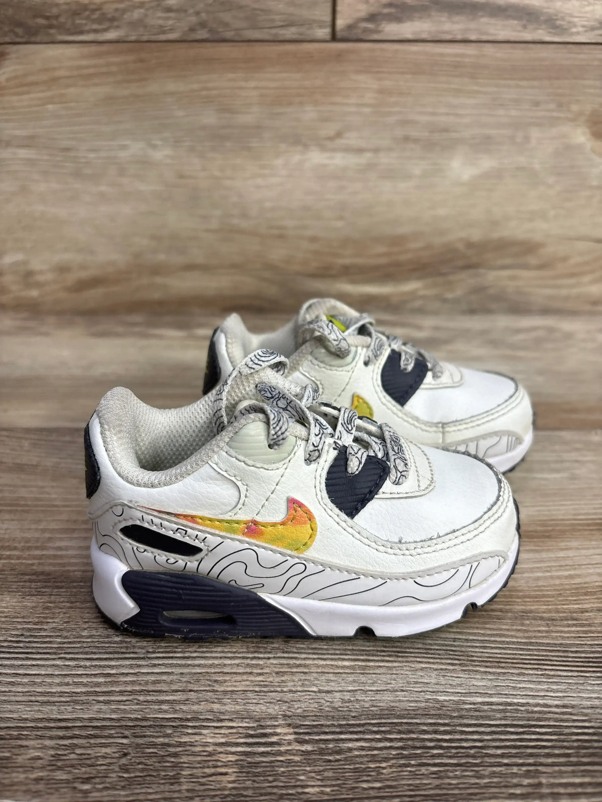 White toddler Nike Air Max sneakers with black accents and yellow-orange swoosh on wooden surface