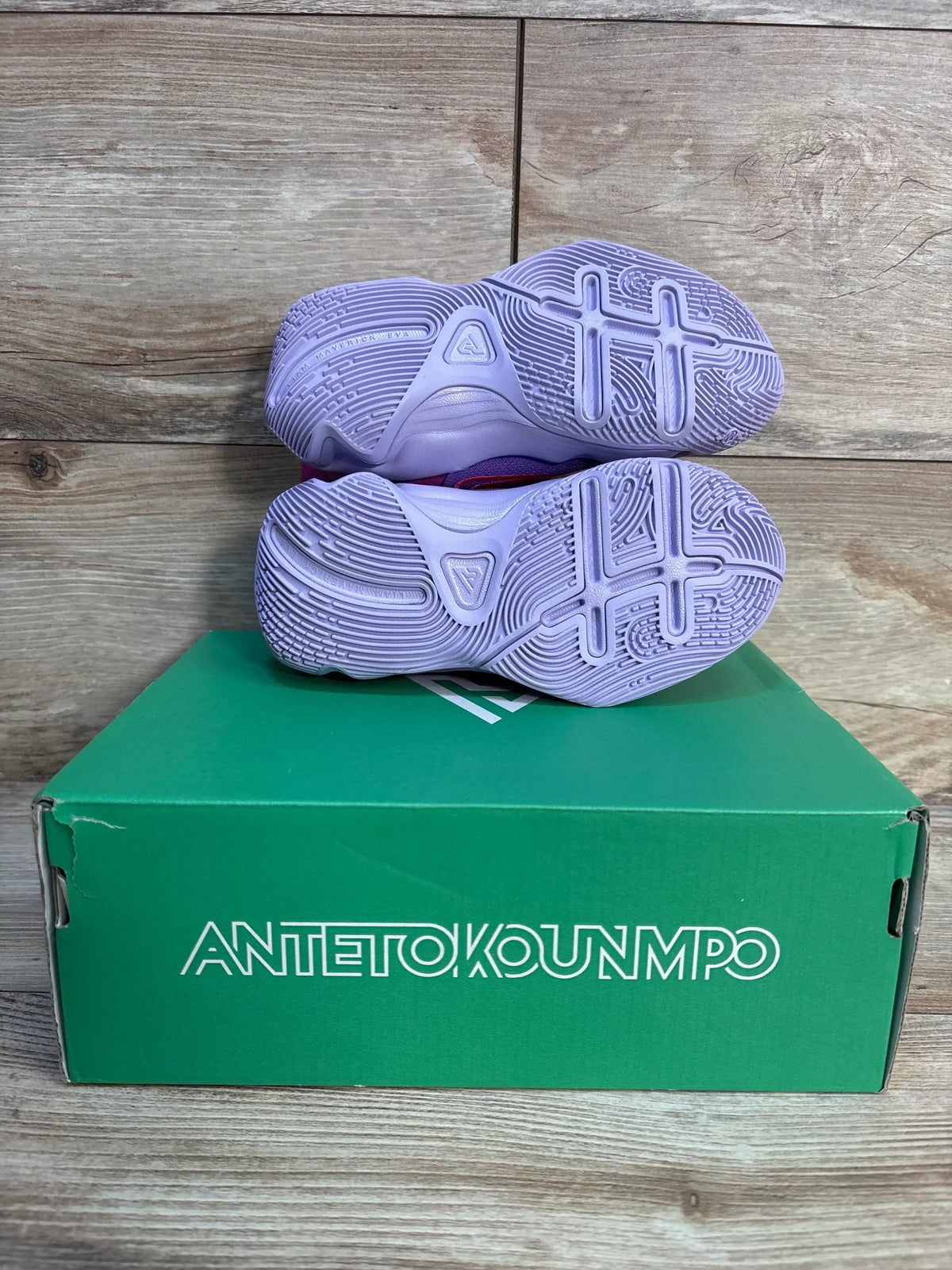 Pair of kids' purple athletic sneakers with patterned soles on green shoe box, wooden floor