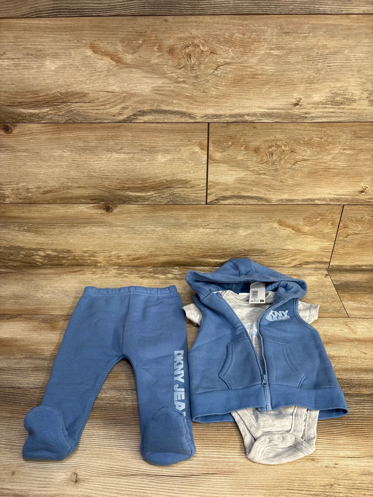 Overhead view of a blue DKNY three-piece baby set showcasing a hooded vest, bodysuit, and footed pants with a wood plank background