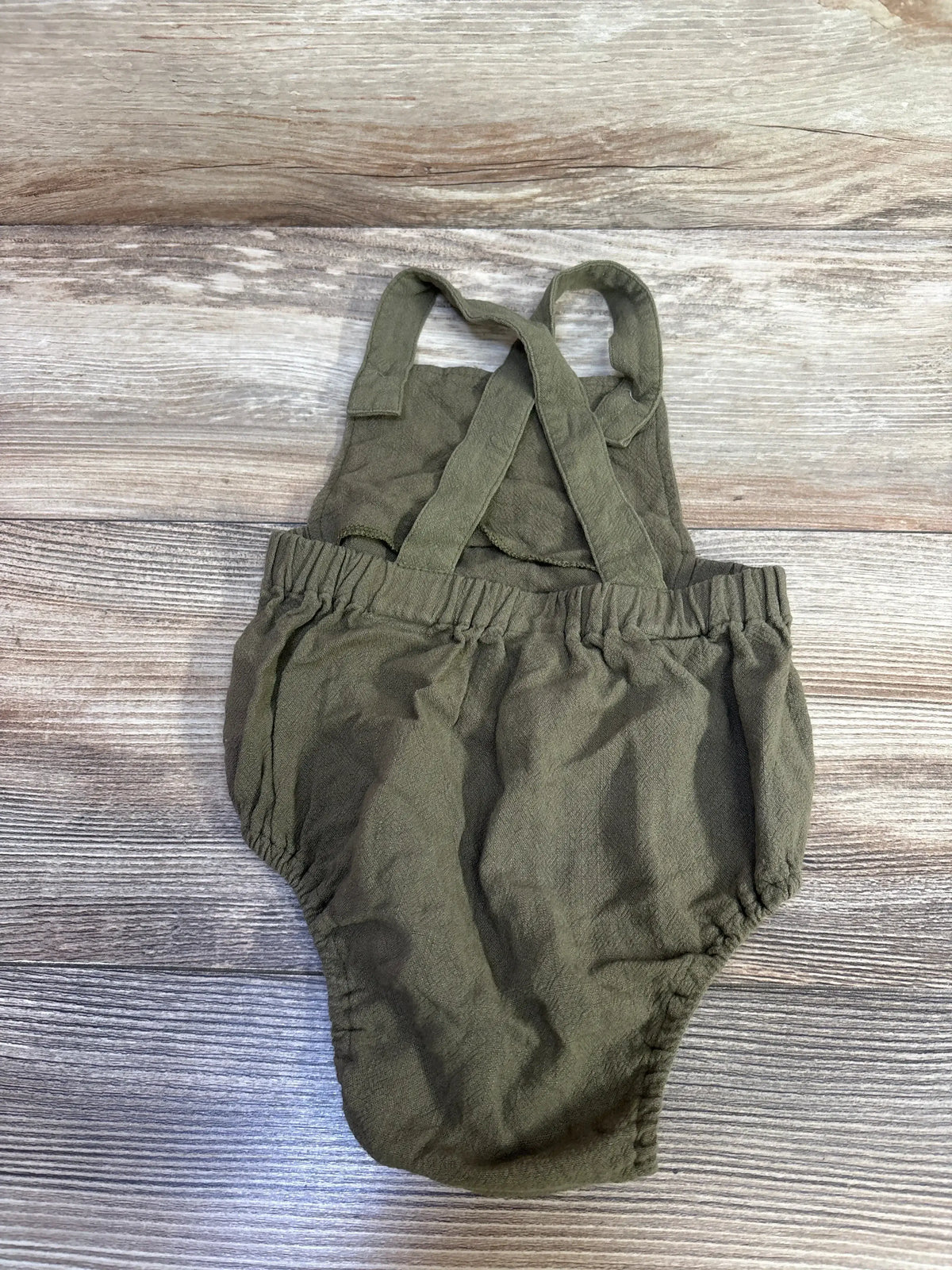 Flat lay of olive green linen baby romper with crossed back straps, elasticized waist and leg openings on wood plank background