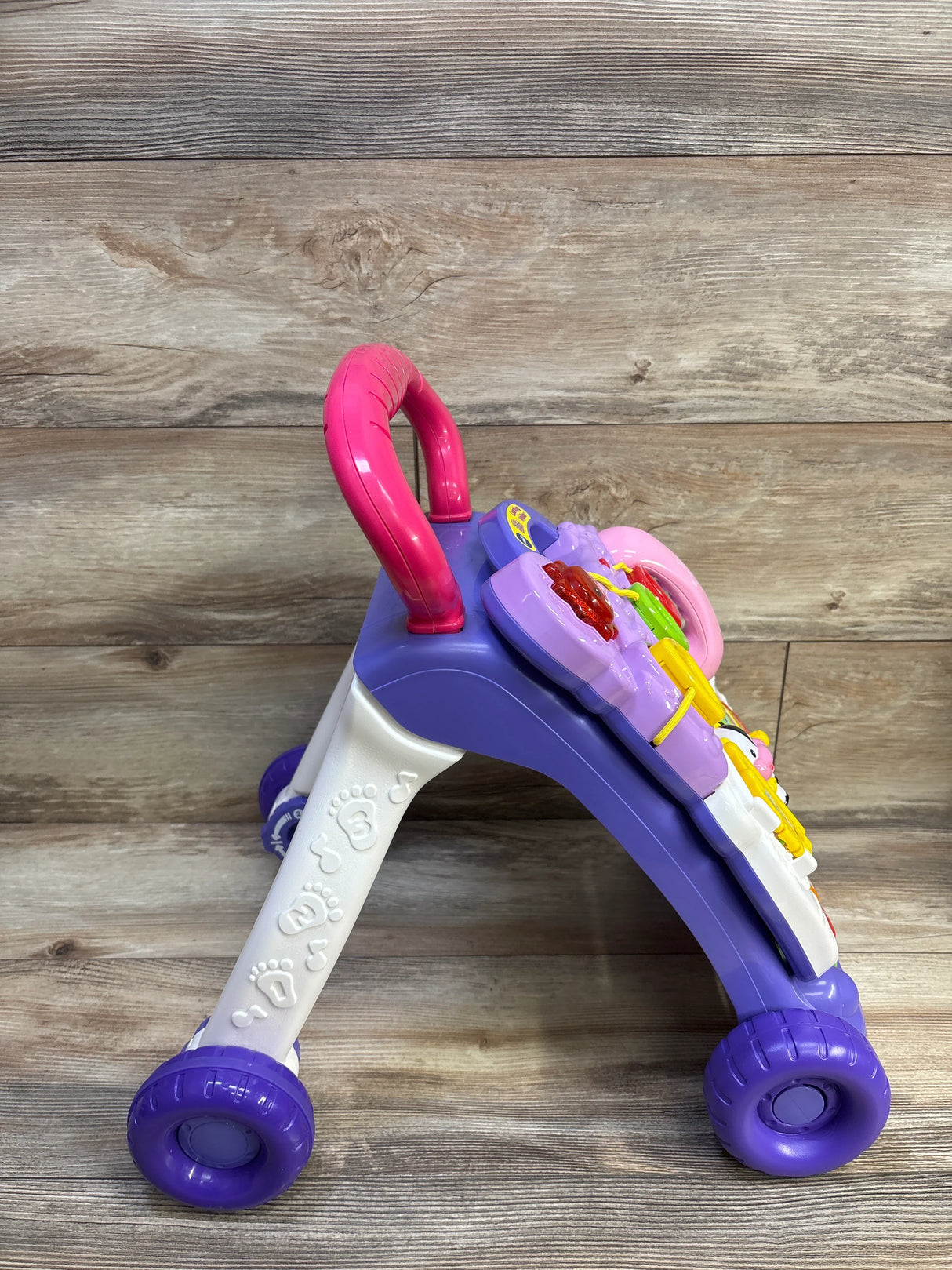 Side view of VTech Sit-to-Stand Learning Walker showing pink handle, purple frame, activity panel and wheels on wood backdrop
