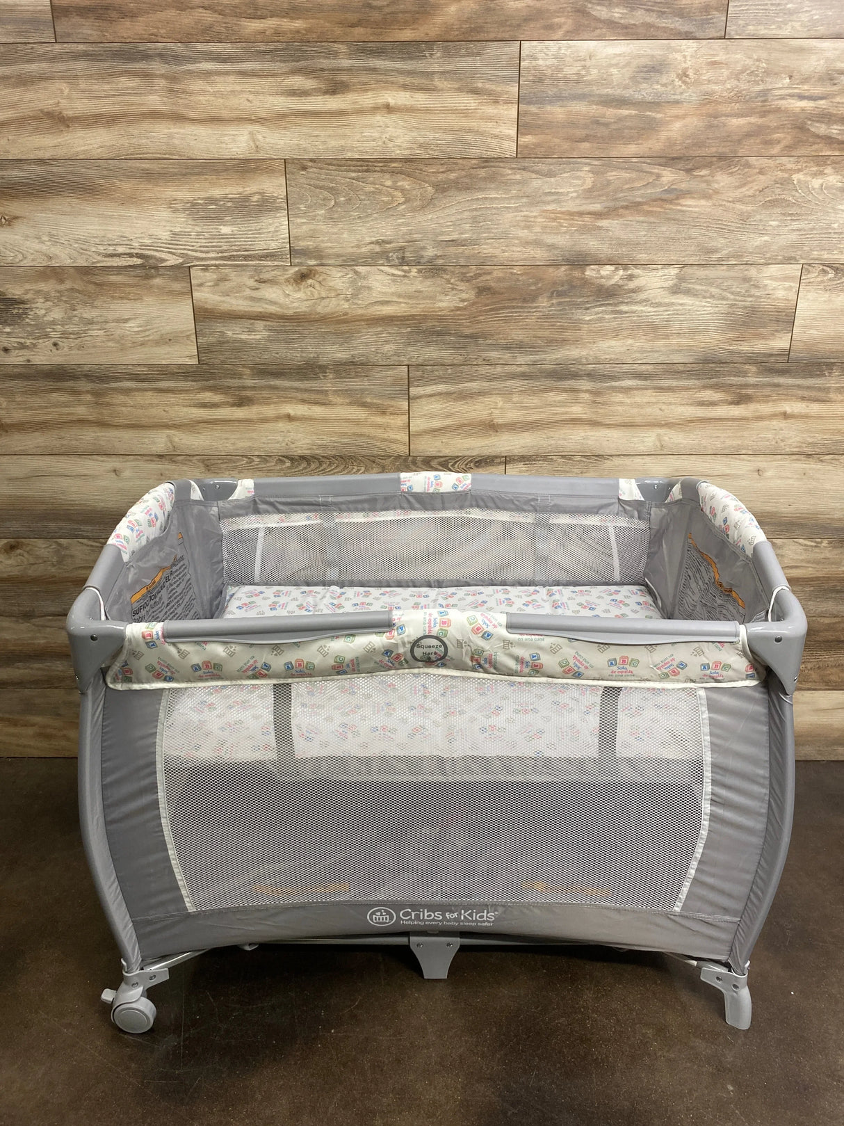 Front view of Cribs for Kids Cribette playard showcasing mesh sides and padded patterned rails against a wood-plank backdrop