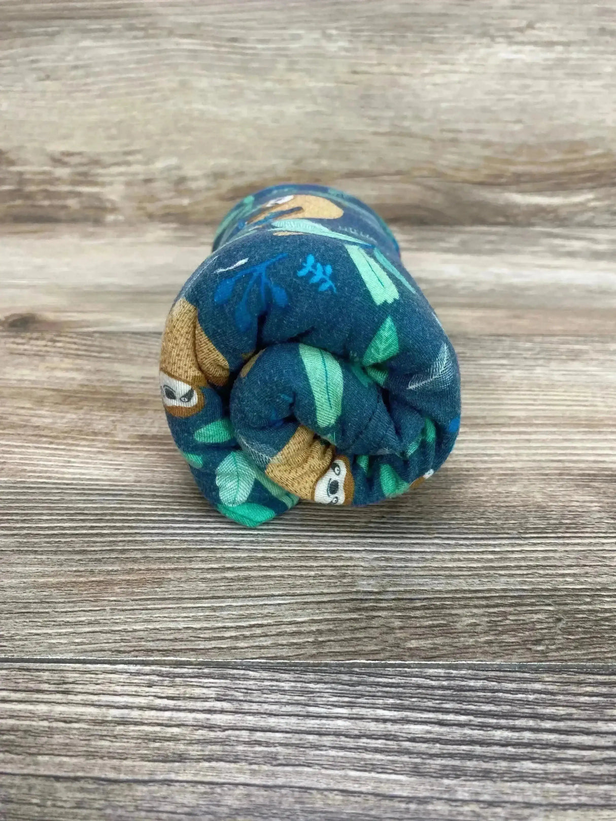 Rolled front view of Milk Snob car seat and nursing cover showing green sloth print on teal fabric against a wood background