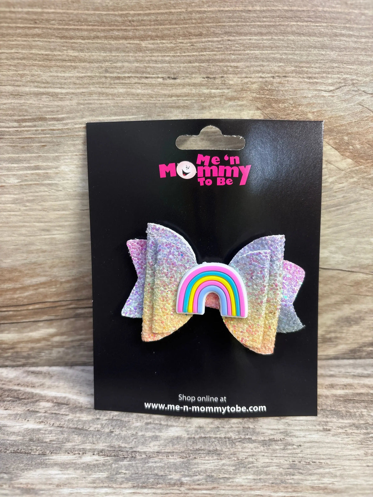 Front view of glitter pastel rainbow hair bow with rainbow applique center on black display card over wood background