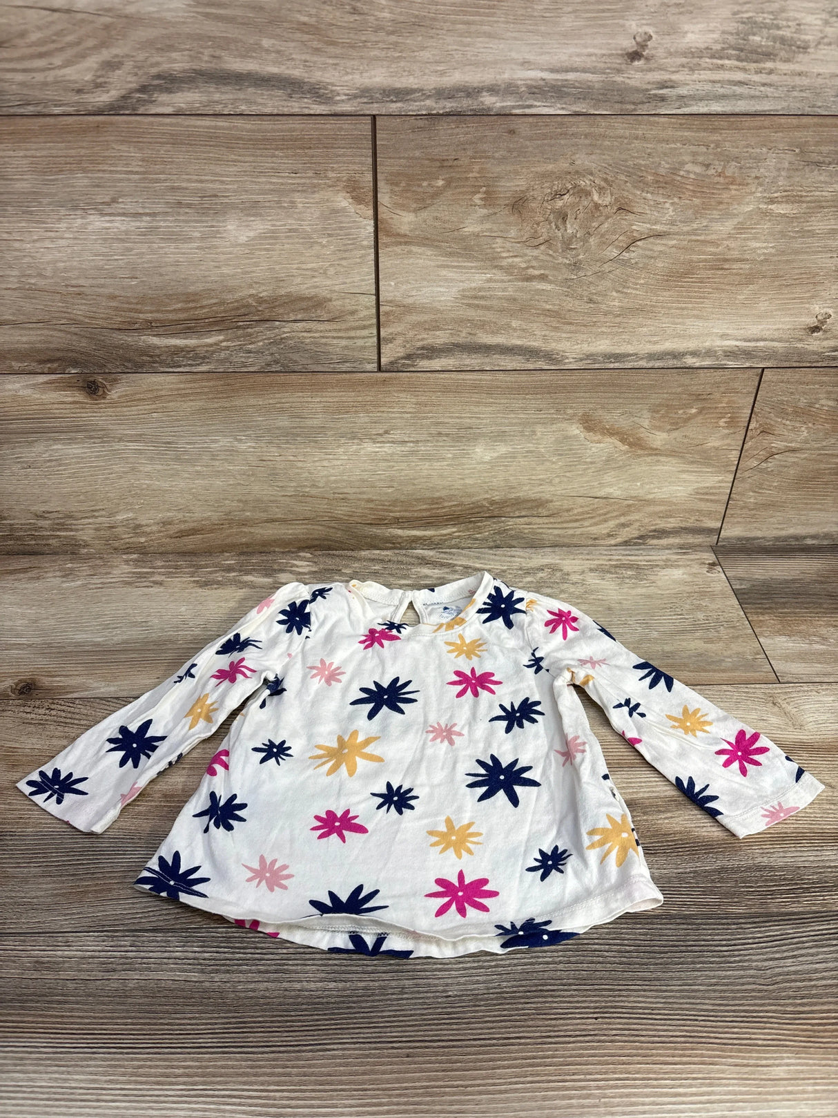 White long-sleeve toddler shirt with pink, navy, and yellow flower patterns on wooden floor
