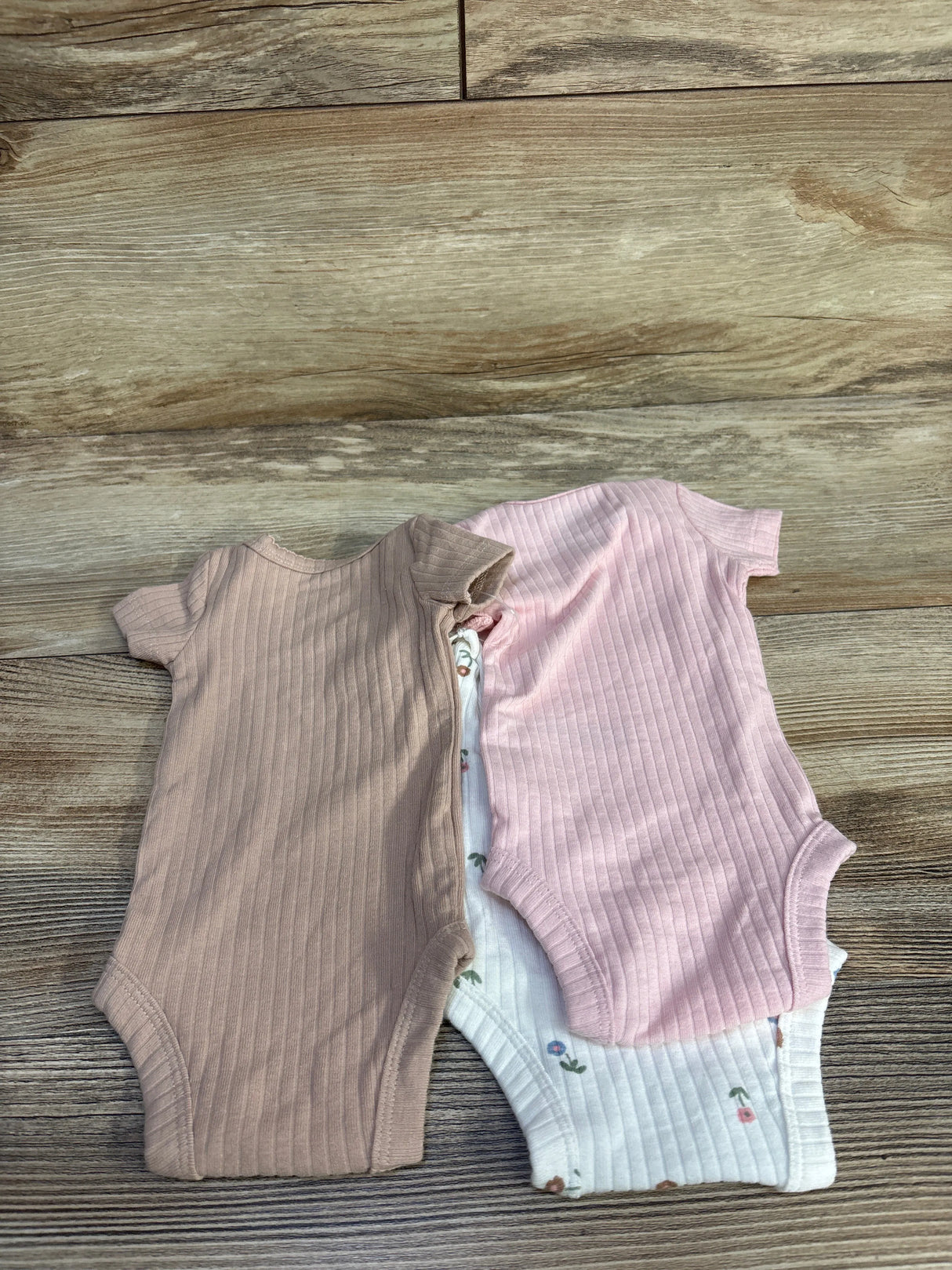 Top-down view of three preemie ribbed short-sleeve bodysuits (dusty pink, light mauve, and white floral) on wood background