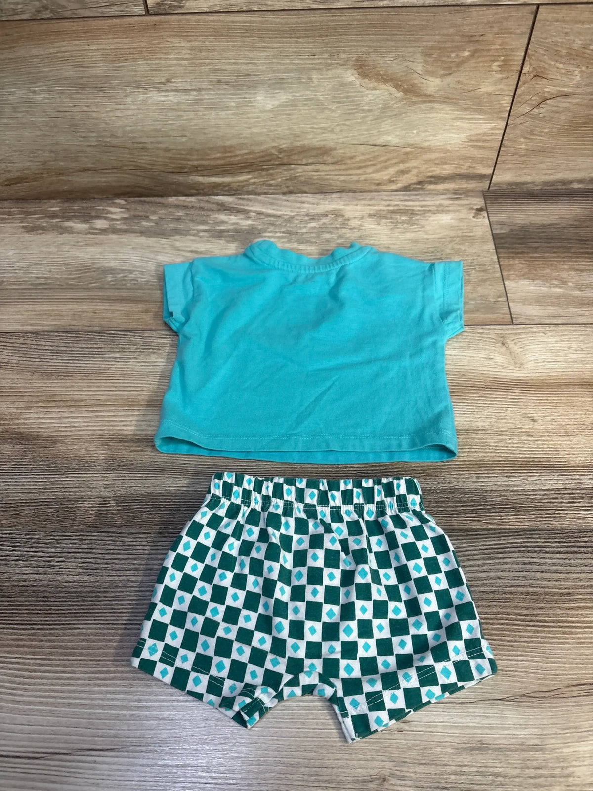 Top-down view of Cat & Jack teal newborn shirt and shorts set showcasing a plain shirt and geometric patterned shorts on a wood floor