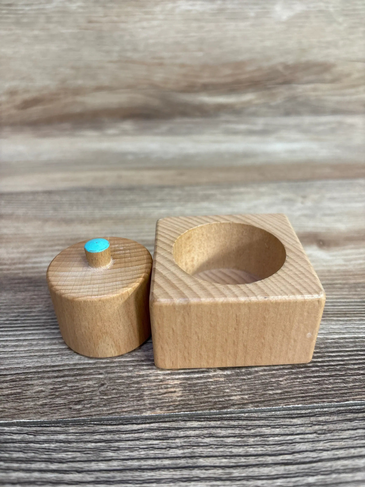 Front view of Lovevery Pincer Puzzle showcasing square wooden box and round piece with blue-topped peg on wooden surface