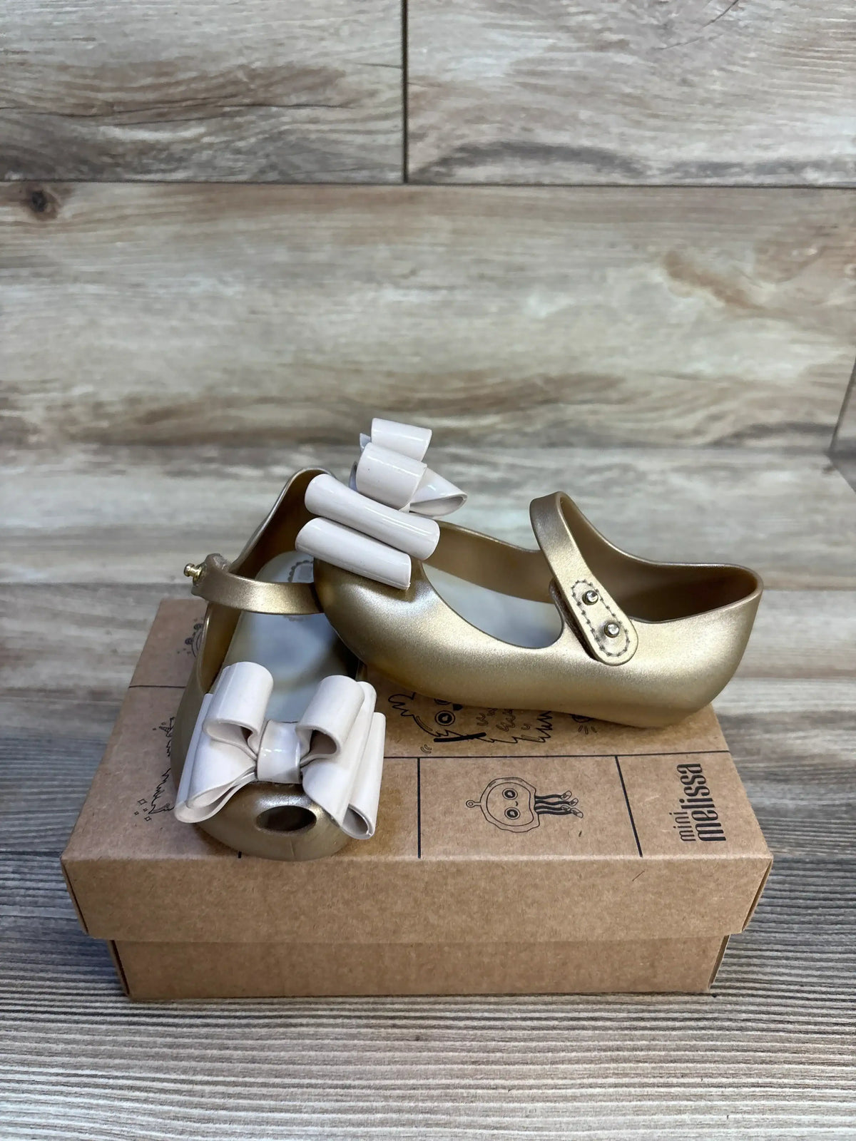 Gold toddler Mary Jane shoes with white bows on cardboard box, wood background