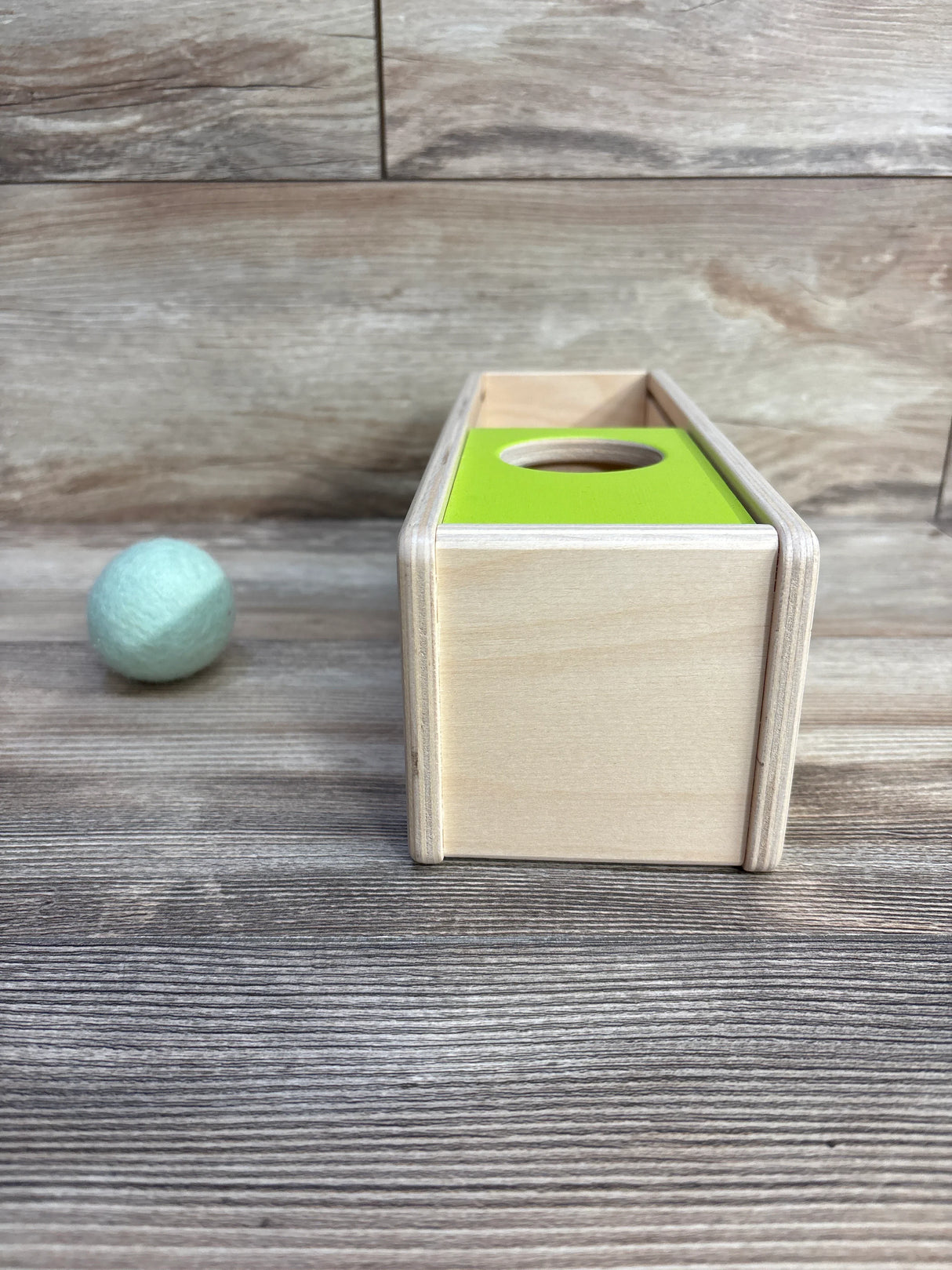 Wooden ball drop toy with green hole and light blue felt ball on wooden floor