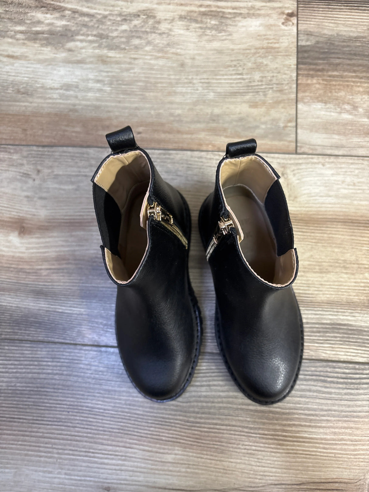Top-down view of a pair of Janie and Jack black Chelsea boots showcasing gold side zippers and elastic gussets on a light wood floor