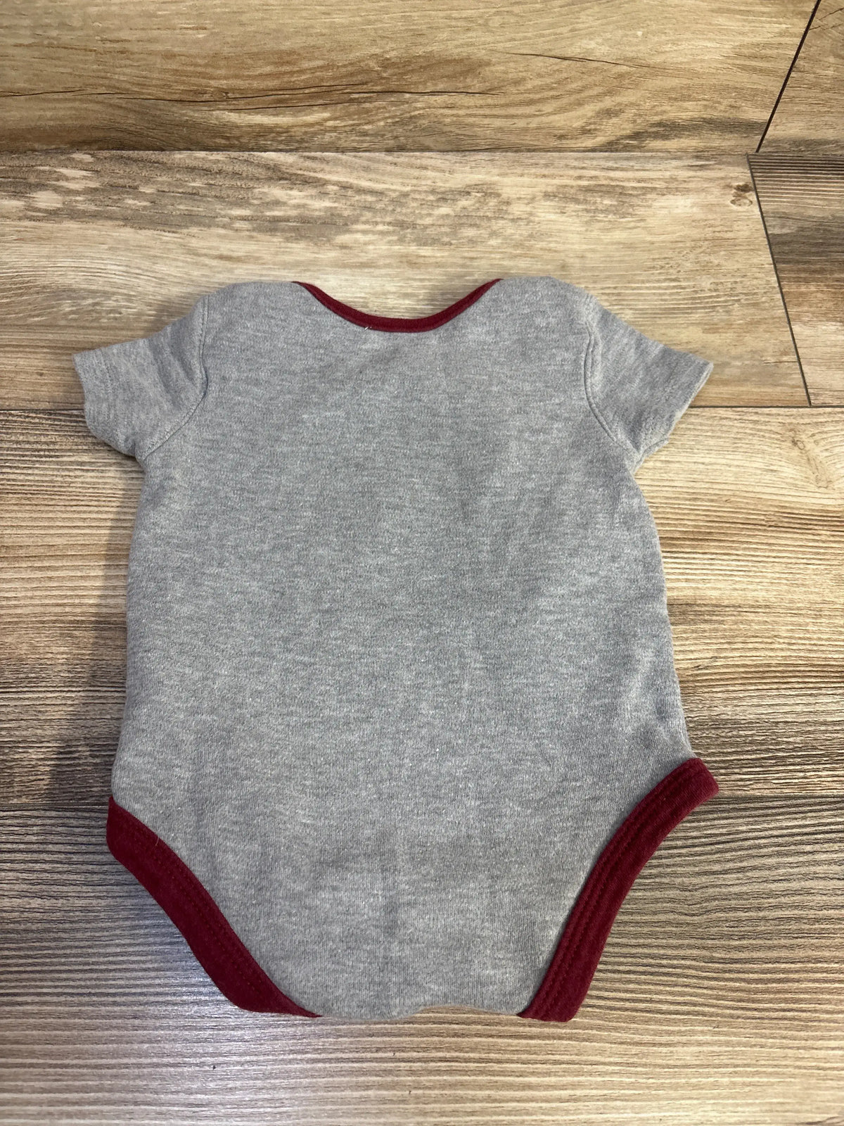 Flat view of NFL Washington Redskins grey bodysuit in size 3/6 months with burgundy trim and short sleeves on a wood floor