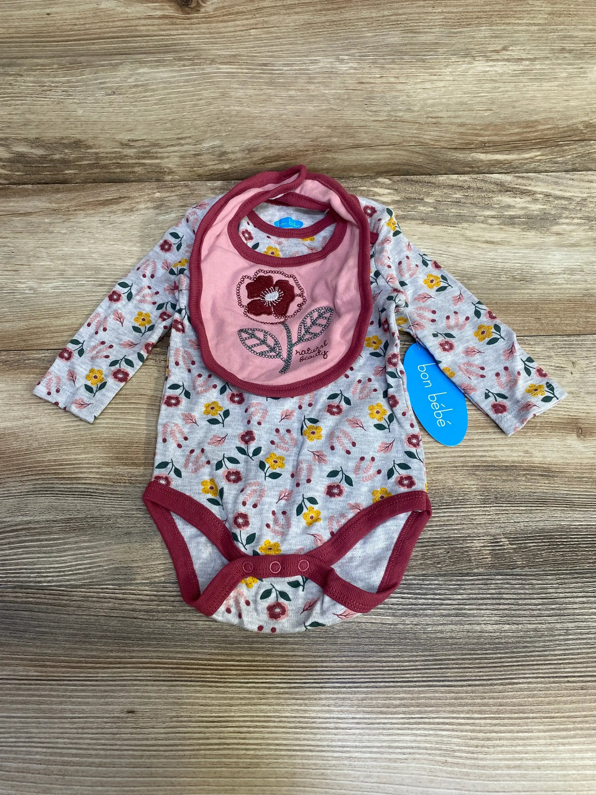 Top-down view of grey floral long-sleeve baby bodysuit with burgundy trim and matching pink embroidered bib, blue bon bebe tag on wood background