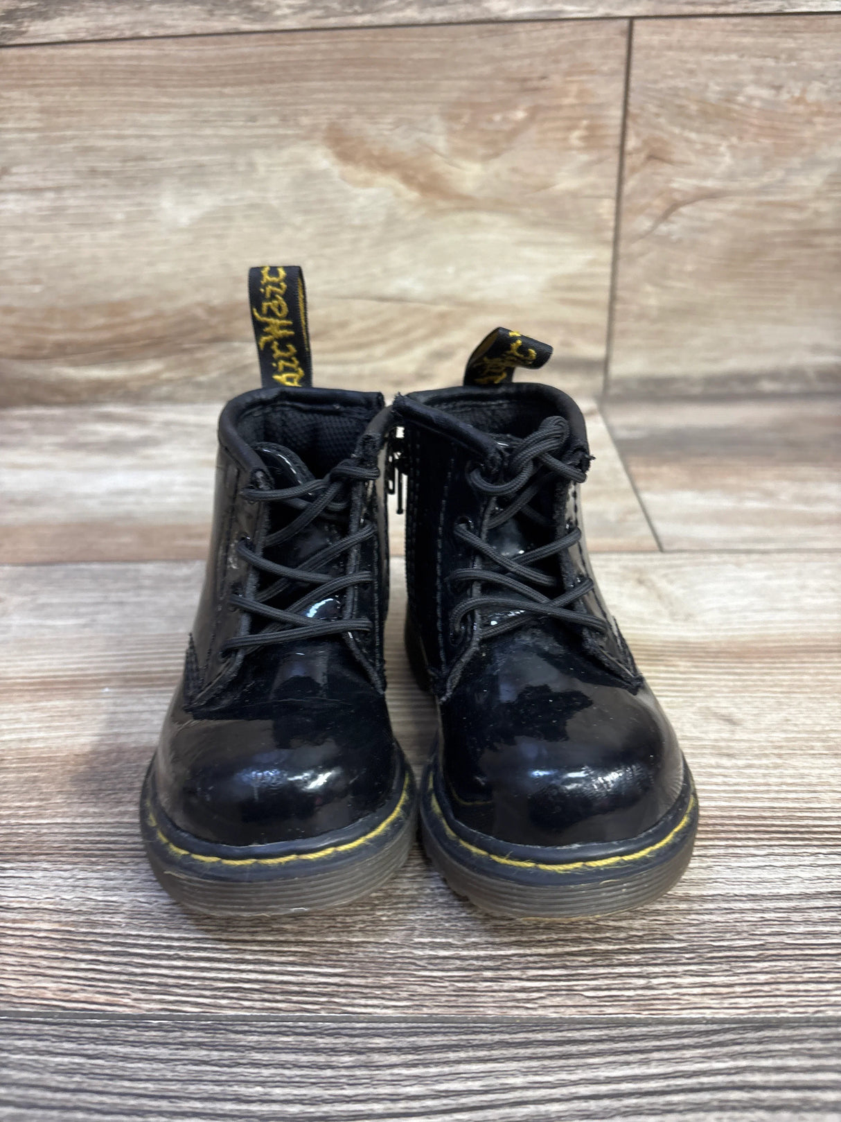 Front view of toddler Dr Martens patent leather boots with glossy black finish and yellow welt stitching on wood floor