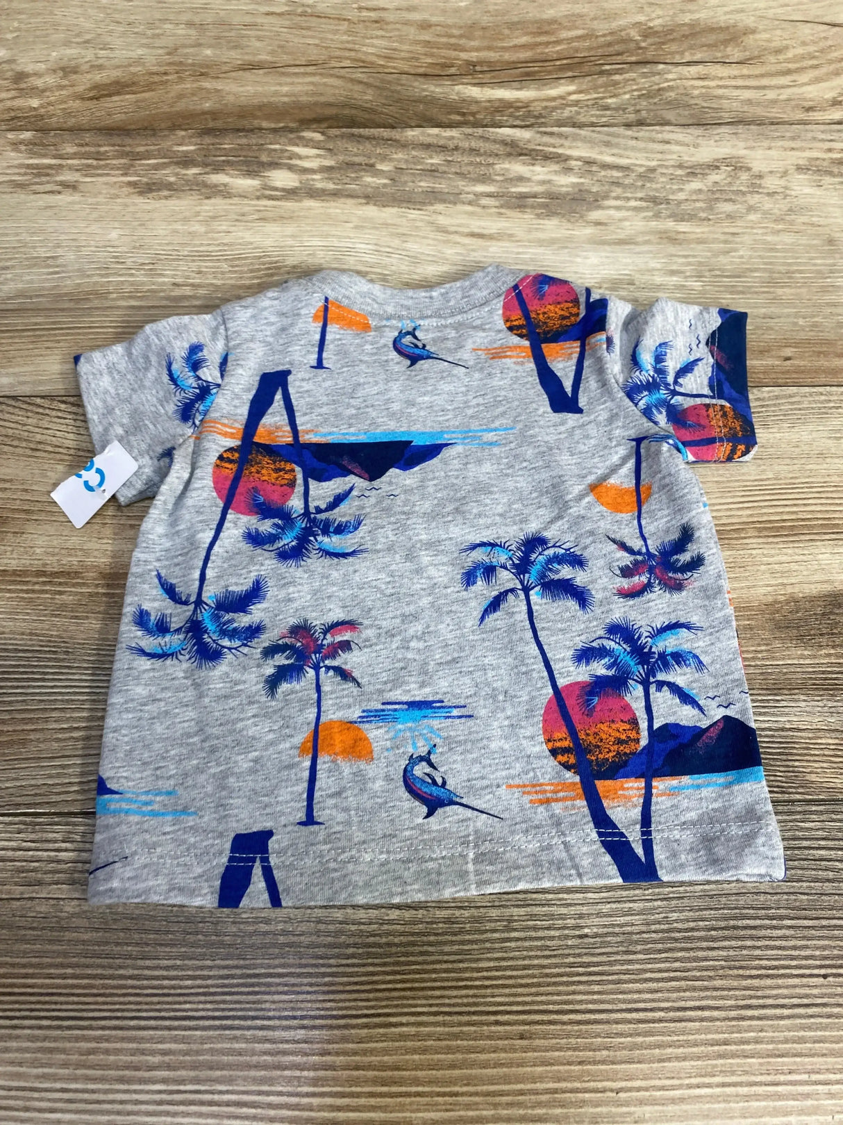 Flat-lay of grey Carter’s baby t-shirt showing blue and orange sunset, palm trees, and marlin print on a wood plank background