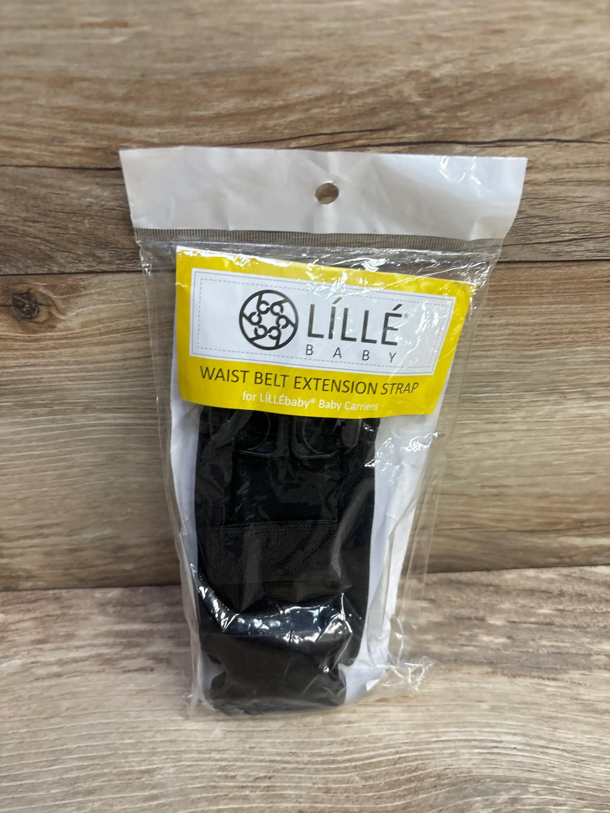Front view of LILLEbaby waist belt extension strap in sealed packaging showing black strap and yellow label on wood surface