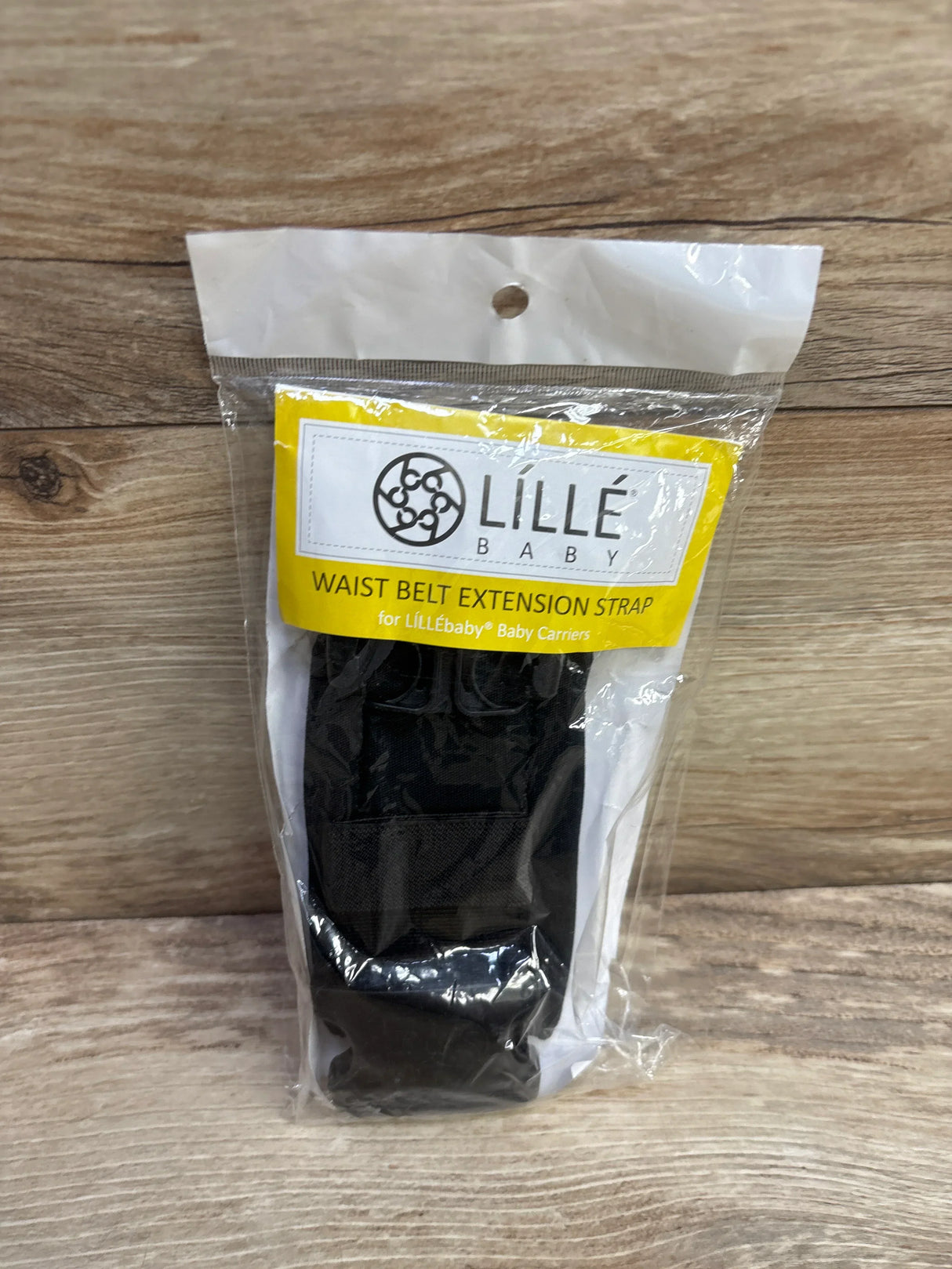 Front view of LILLEbaby waist belt extension strap in sealed packaging showing black strap and yellow label on wood surface