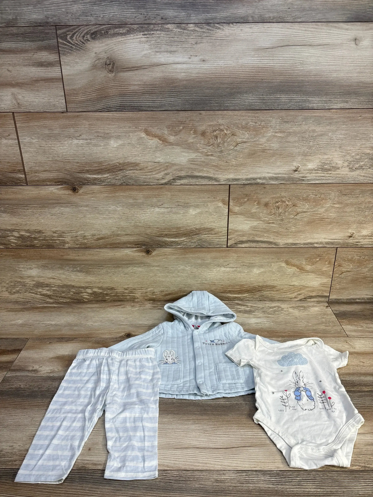 Flat lay of Peter Rabbit 3pc Hooded Jacket Bodysuit and Pants Set in blue stripes with rabbit graphic on wood plank floor