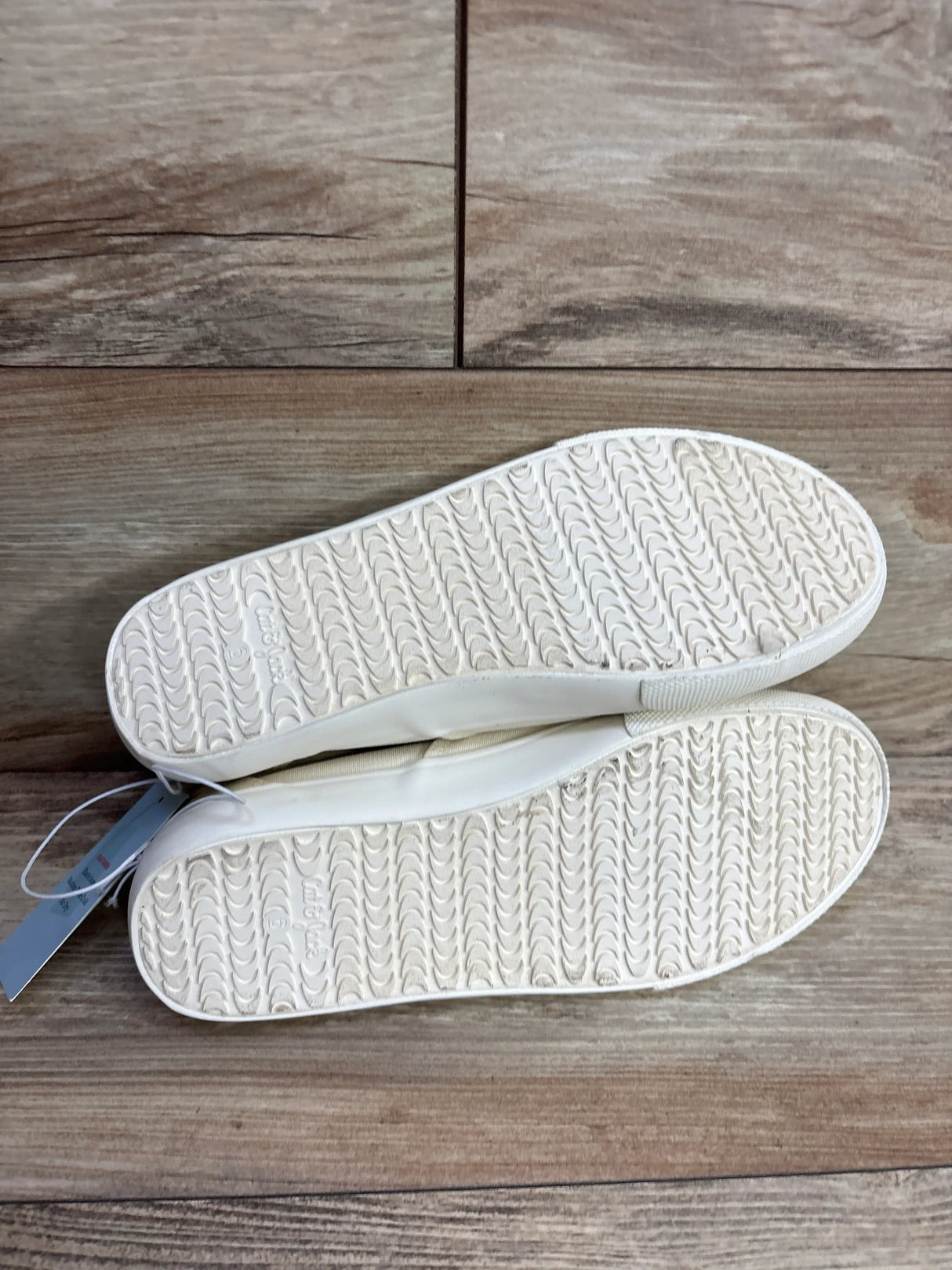 Pair of white kids' slip-on shoes with textured rubber soles on wood floor
