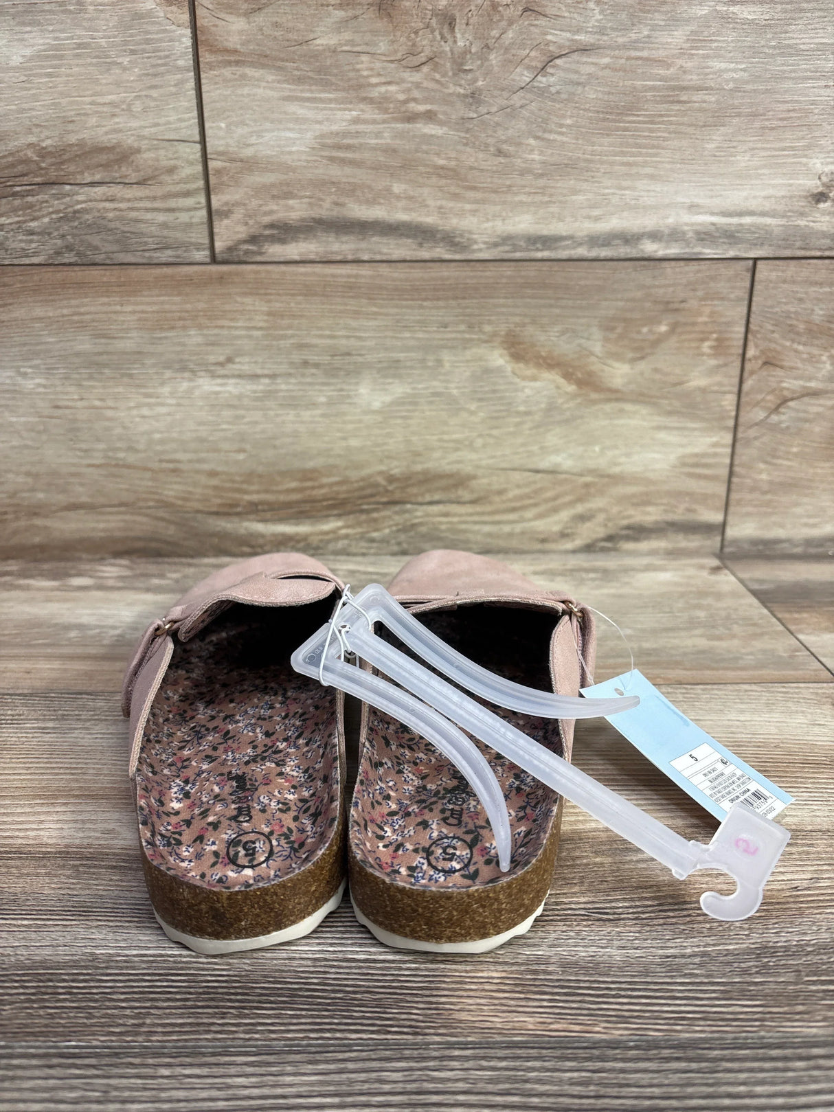 Pair of pink kid's slippers with floral insoles and cork soles on wooden floor, tag and plastic hanger attached