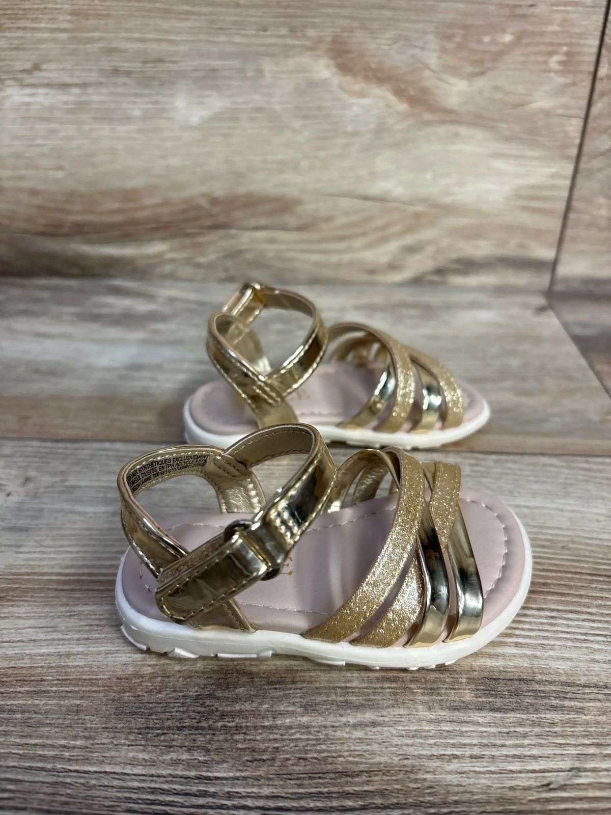 Three-quarter view of Children’s Place gold glitter and metallic sandals with ankle buckles and white soles on a wood surface