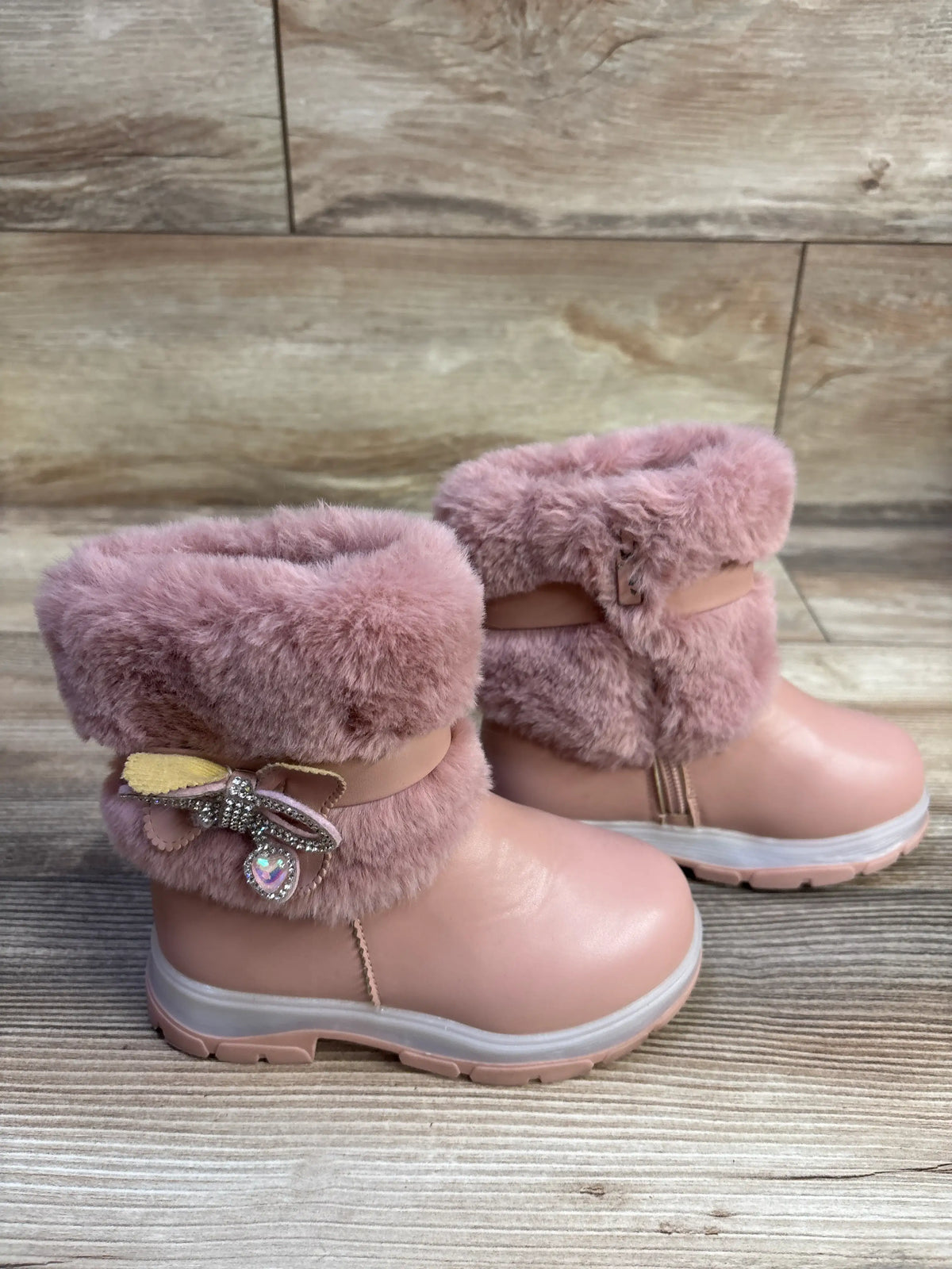 Front three-quarter view of pink Belladia faux-fur boots showcasing jeweled bows and plush fur cuffs on a wood-look floor