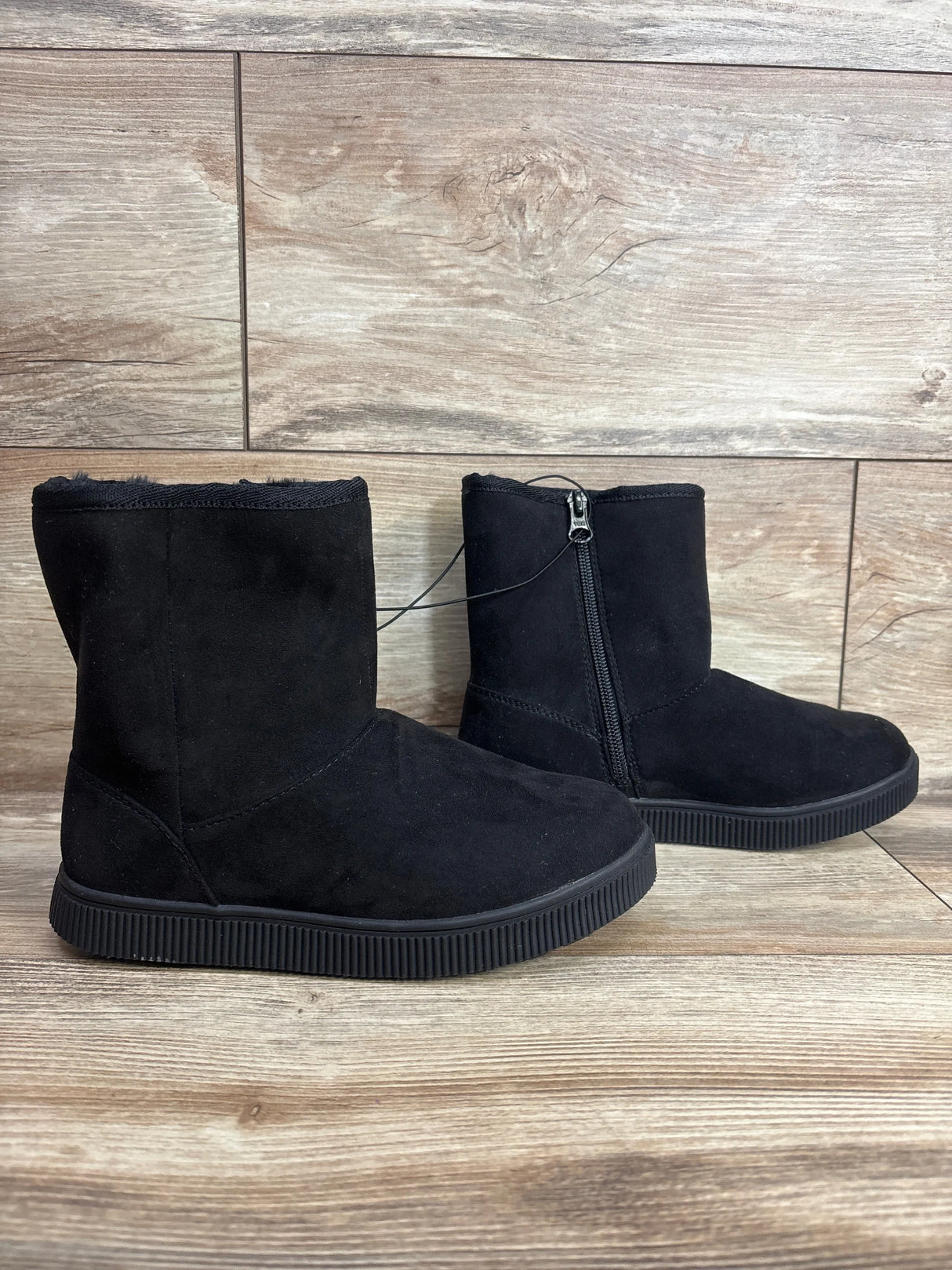 Three-quarter view of black suede kids winter boots with side zipper and faux fur lining on wood plank floor