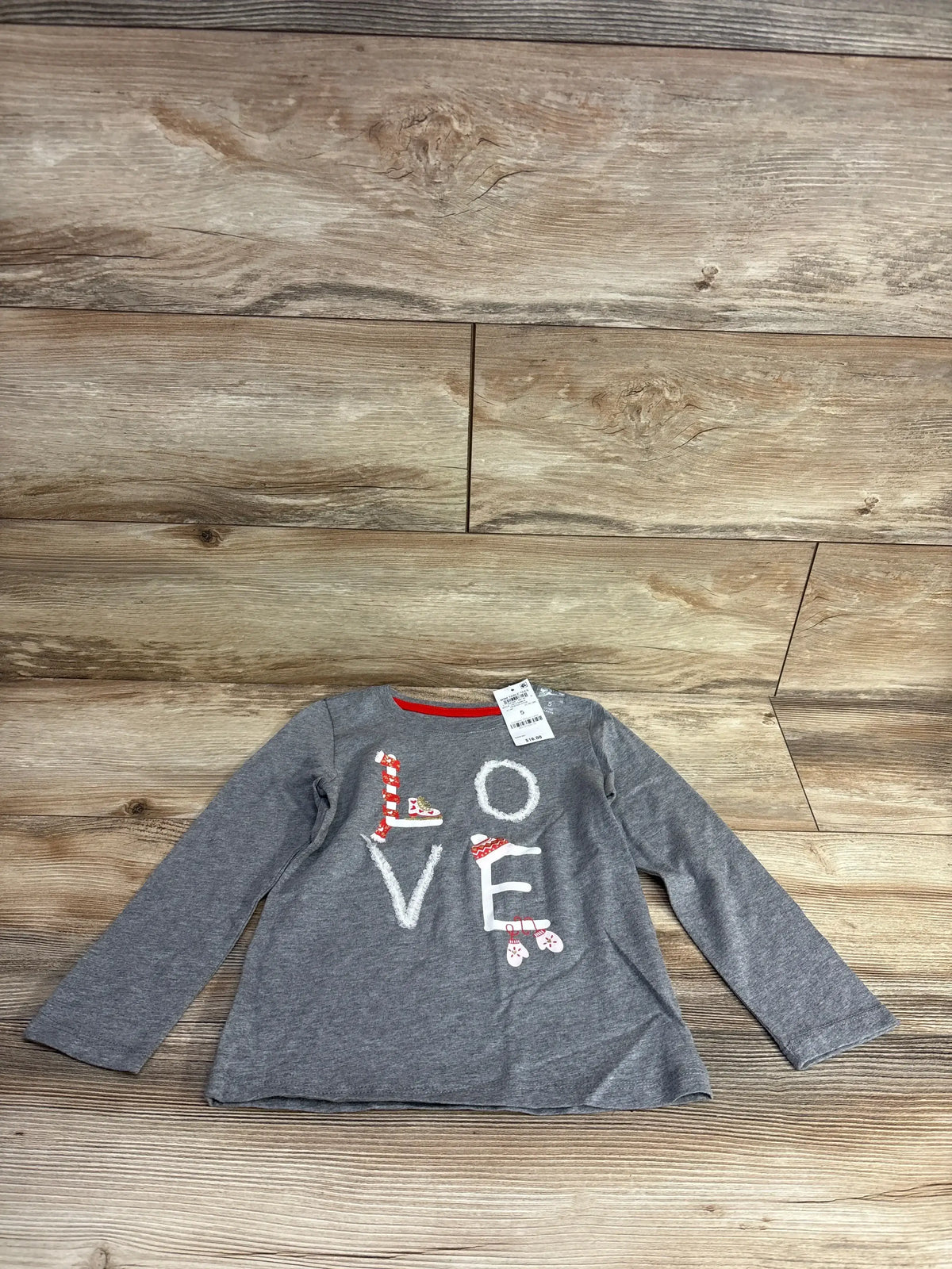 Gray kids long sleeve shirt with 'LOVE' graphic, candy cane, ice skates, and mittens on wood floor