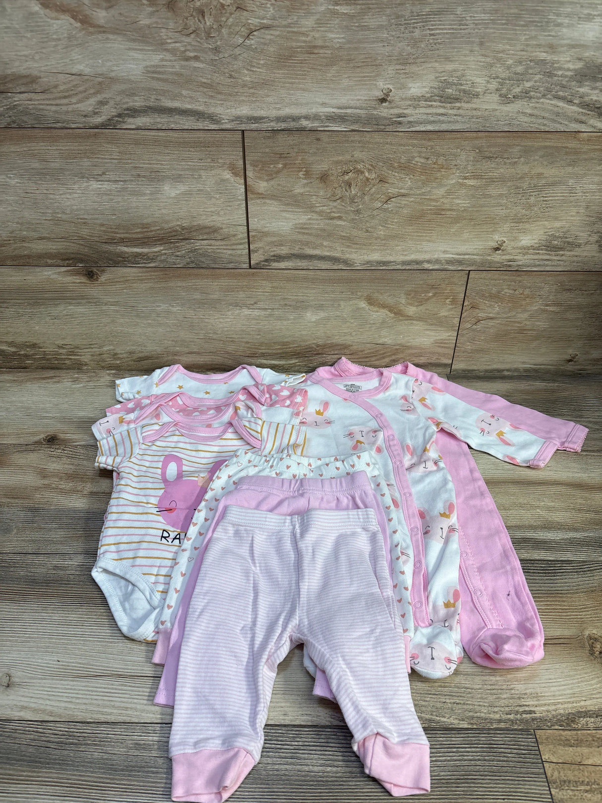 Flat lay of MAKIMAMA 9-piece layette gift set in pink showing assorted bodysuits, footed sleepers, and pants on wood floor