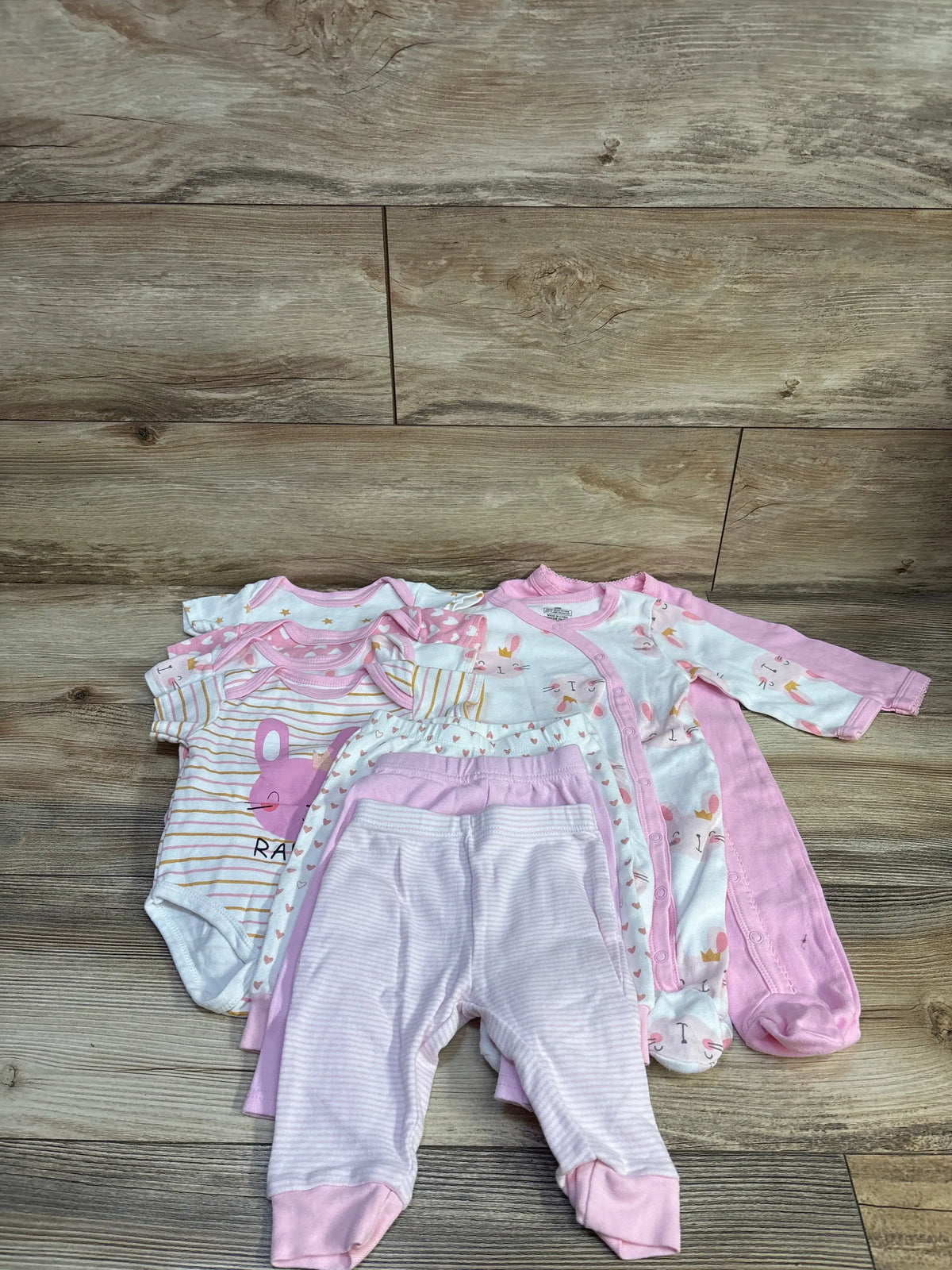 Flat lay of MAKIMAMA 9-piece layette gift set in pink showing assorted bodysuits, footed sleepers, and pants on wood floor