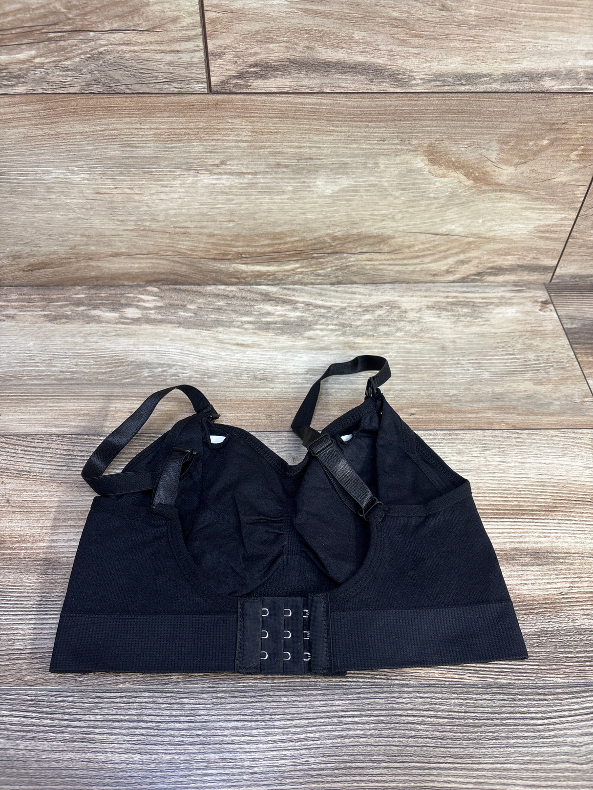 Top-down view of black nursing bra with adjustable straps and nursing clips, showing three-row hook-and-eye back on wood floor background