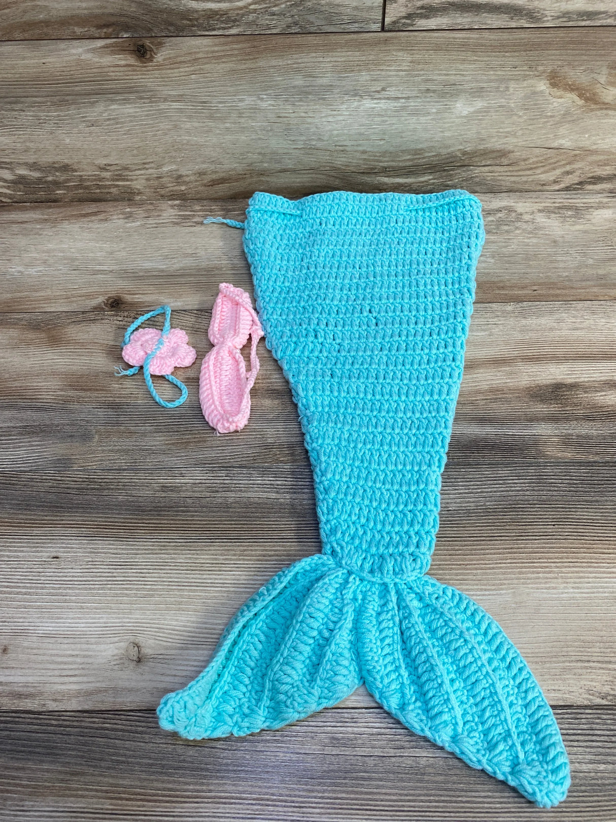 Top-down view of blue crochet mermaid tail with flared fin, pink crochet top and headband on wood background, size 18 months to 2T