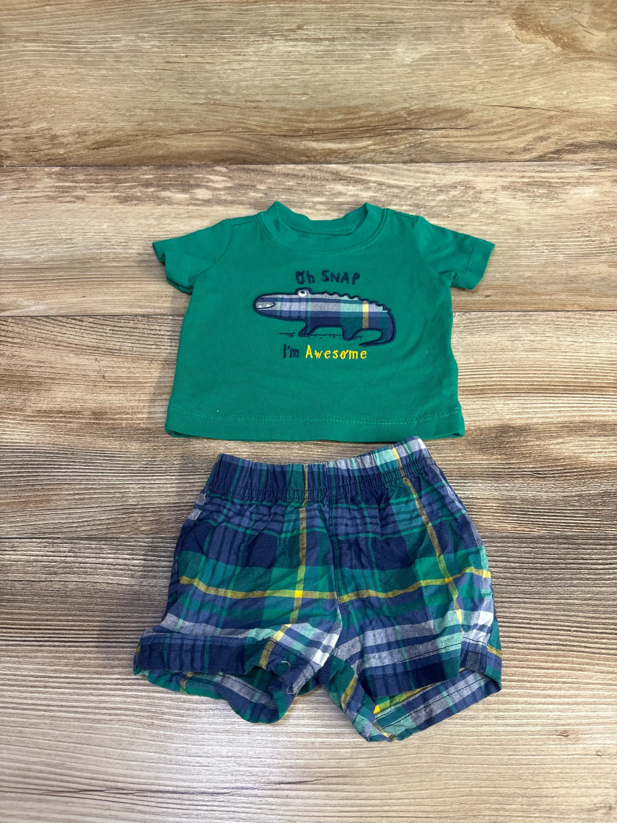 Top-down view of Carter’s newborn shirt and shorts set showing green shirt with alligator graphic and plaid shorts on wood