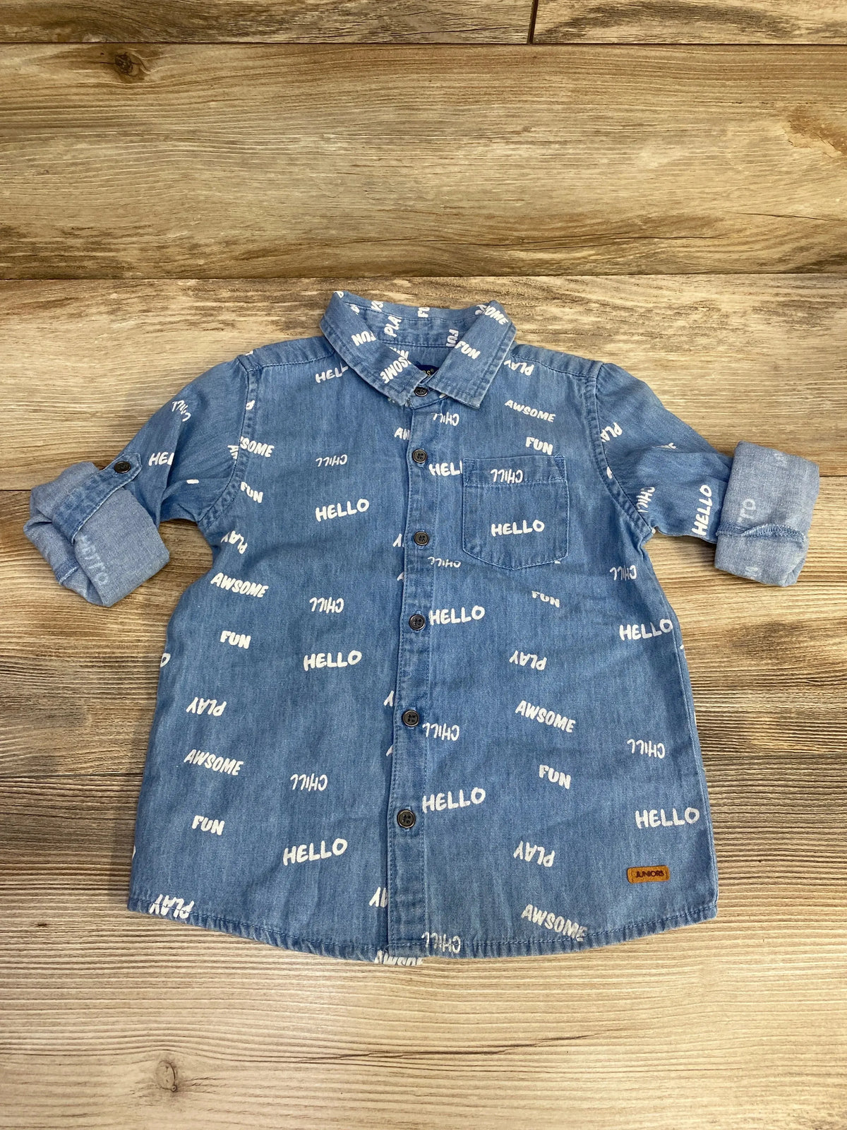 Flat lay of blue chambray button-up shirt showing all-over white HELLO and play print, chest pocket and rolled sleeves on wood background